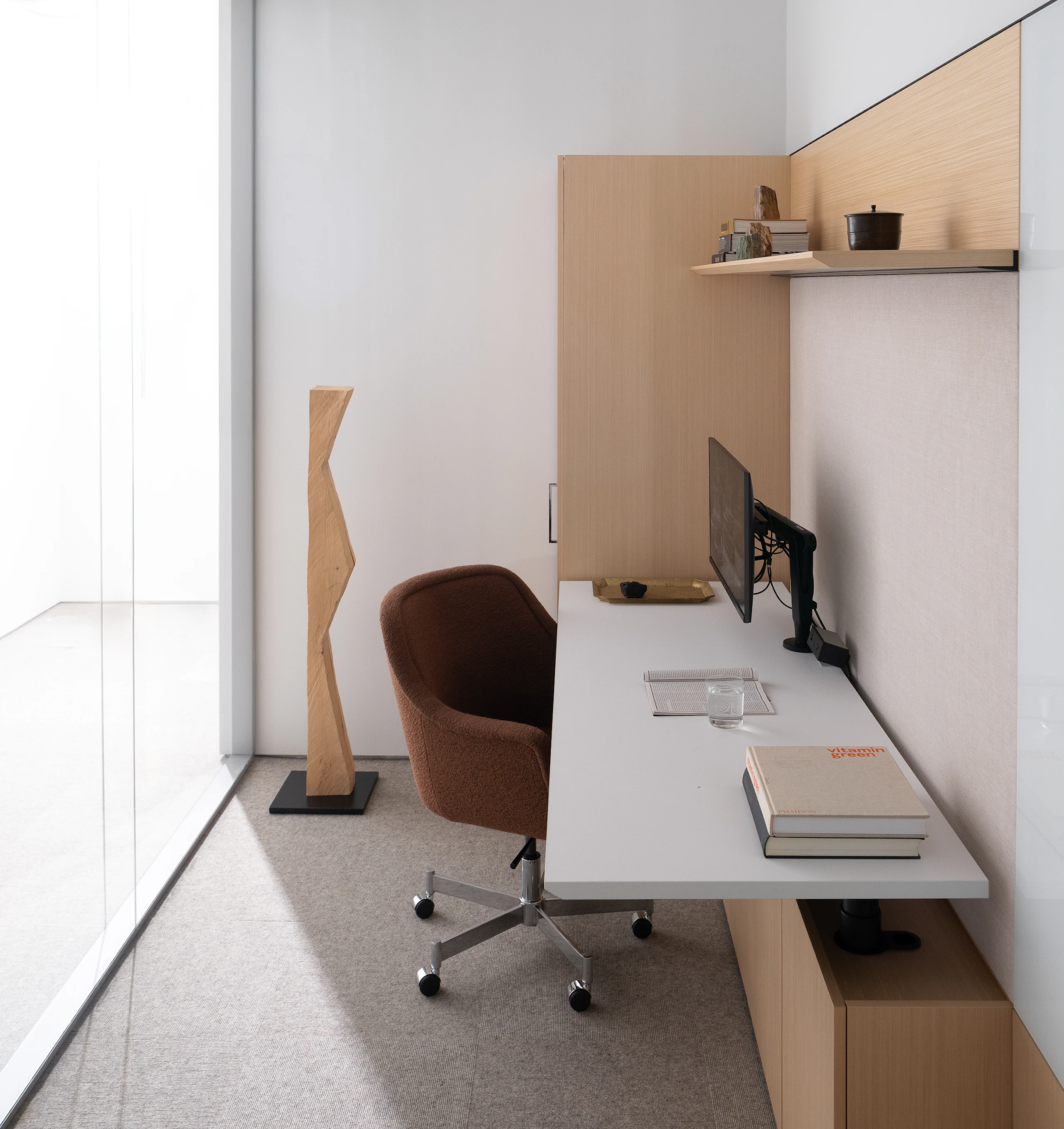 A Geiger One Private Office in Recograin Quarter Cut Rima Oak with a white height-adjustable work surface and a Bumper Chair designed by Ward Bennett upholstered with a brown textile, viewed from the side.