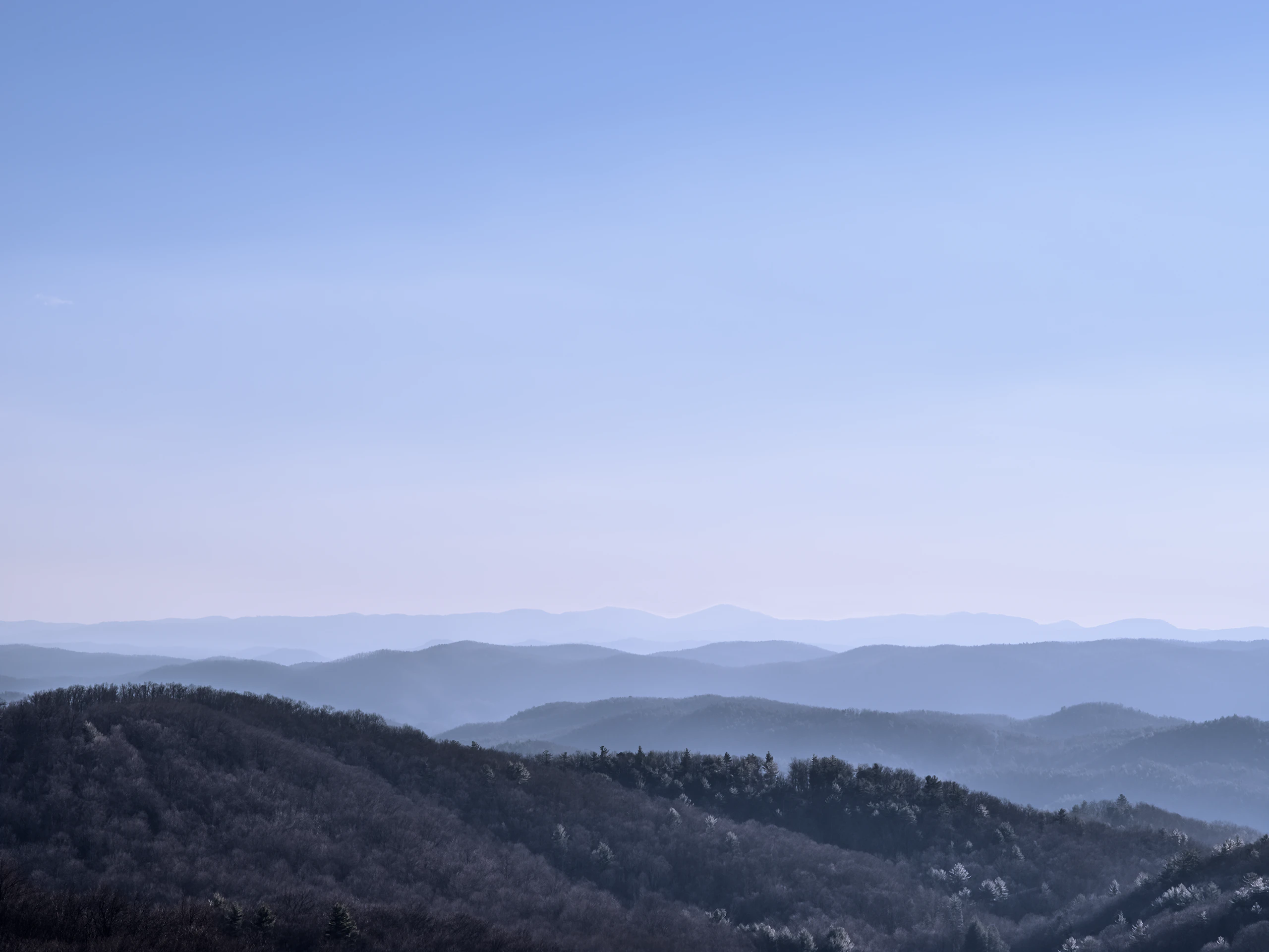 Clear blue sky among layers of mountains covered in trees.