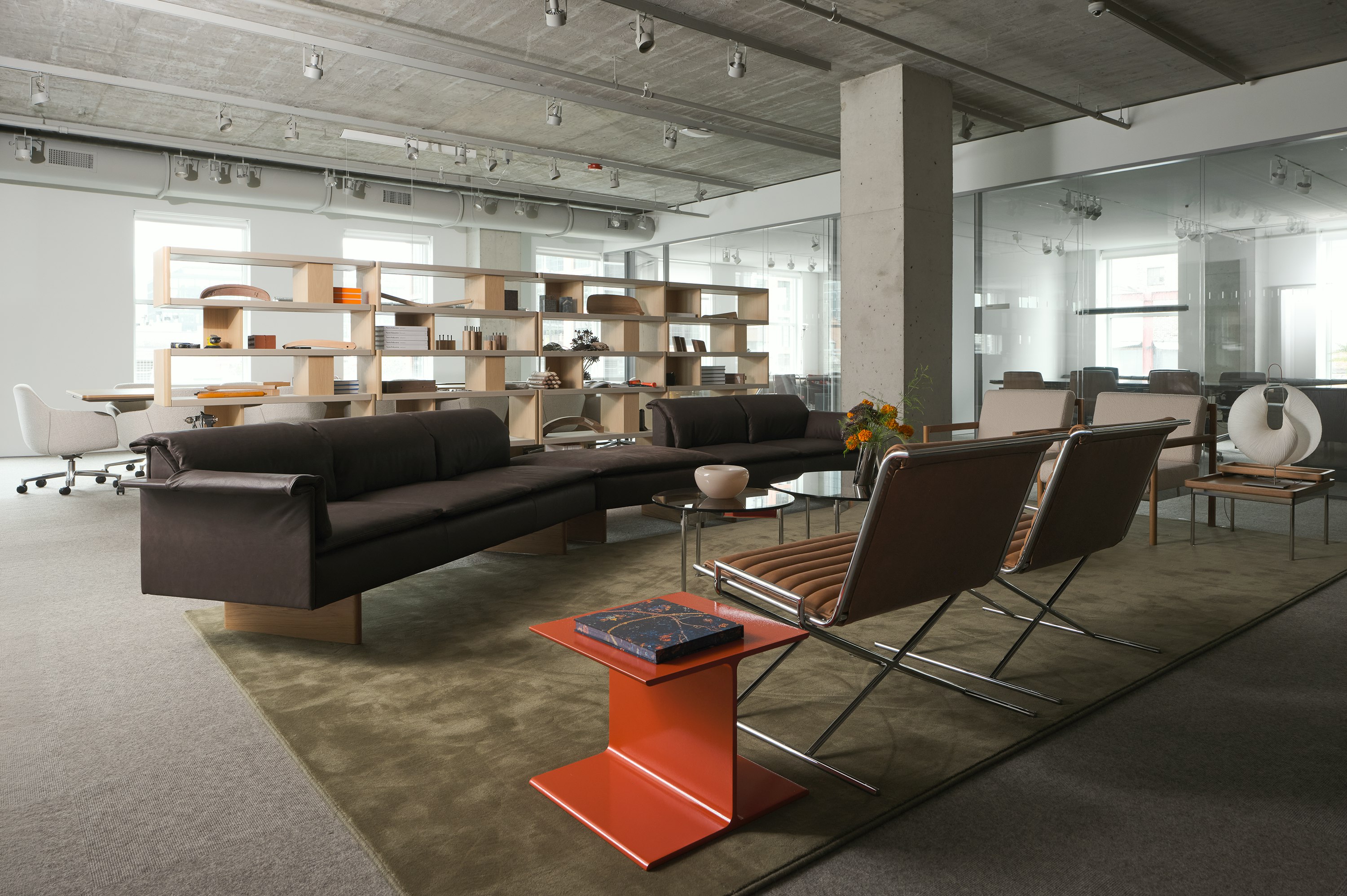 A lounge setting featuring an orange I Beam Side Table, Mantle Sectional Sofa, two Sled Chairs, two round Claw Tables, and two Brabo Club Chairs, and New Canaan Bookshelves in the background, viewed at an angle in a showroom setting.