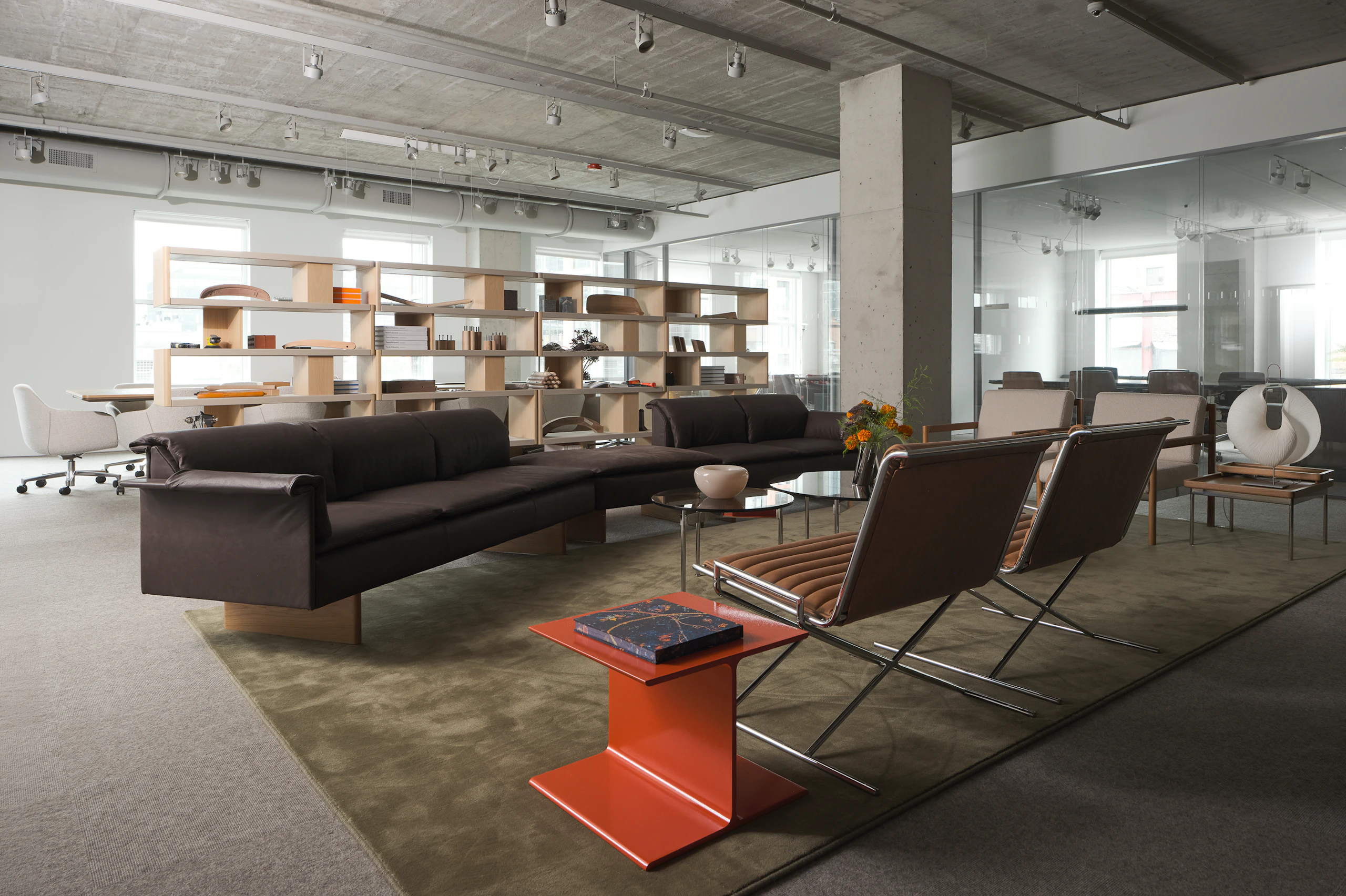 A lounge setting featuring an orange I Beam Side Table, Mantle Sectional Sofa, two Sled Chairs, two round Claw Tables, and two Brabo Club Chairs, and New Canaan Bookshelves in the background, viewed at an angle in a showroom setting.