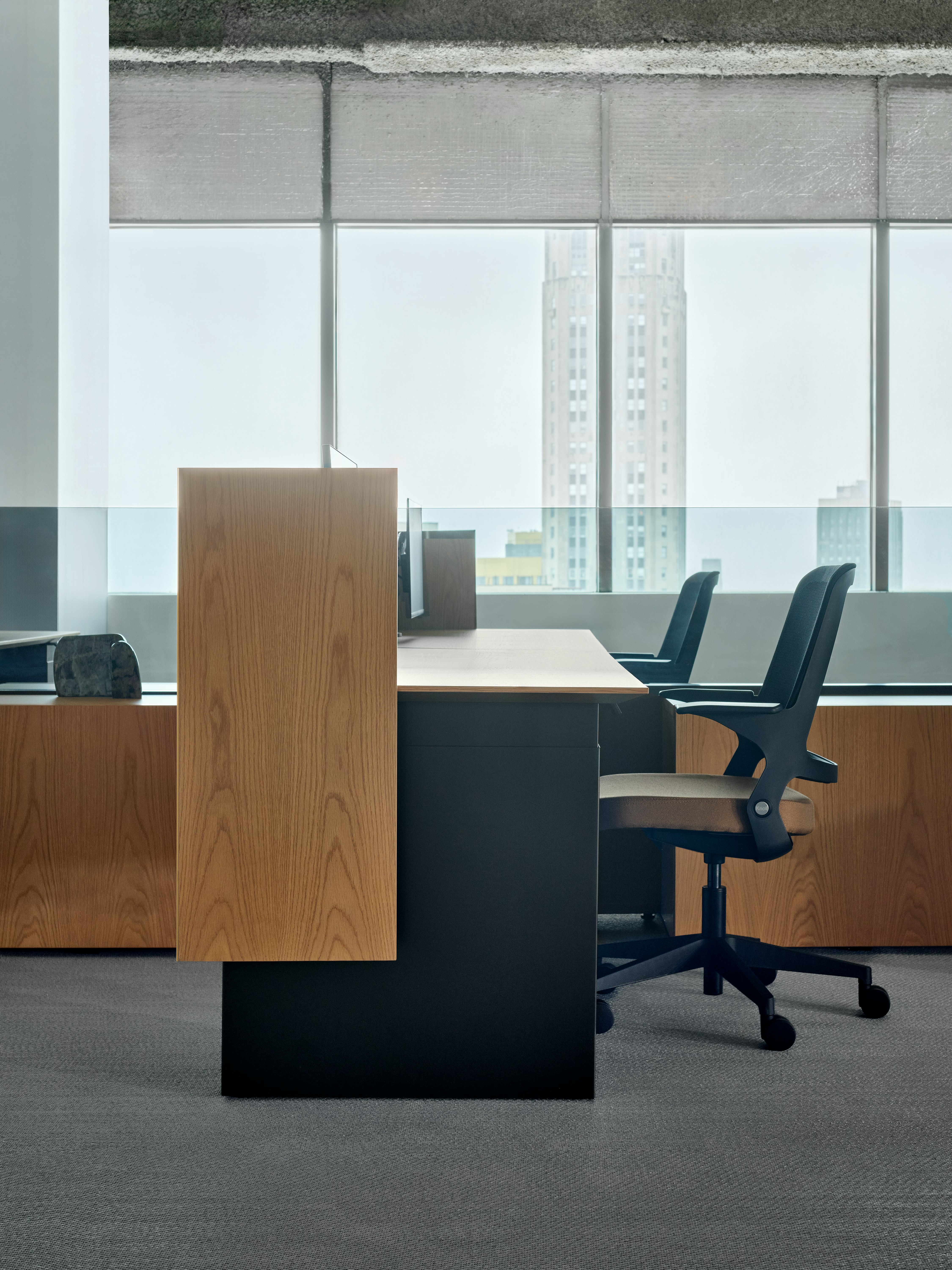Two desks with table mounted screens are viewed from the side. Each desk has a work chair in front of it and the desks are separated by a glass screen add on.