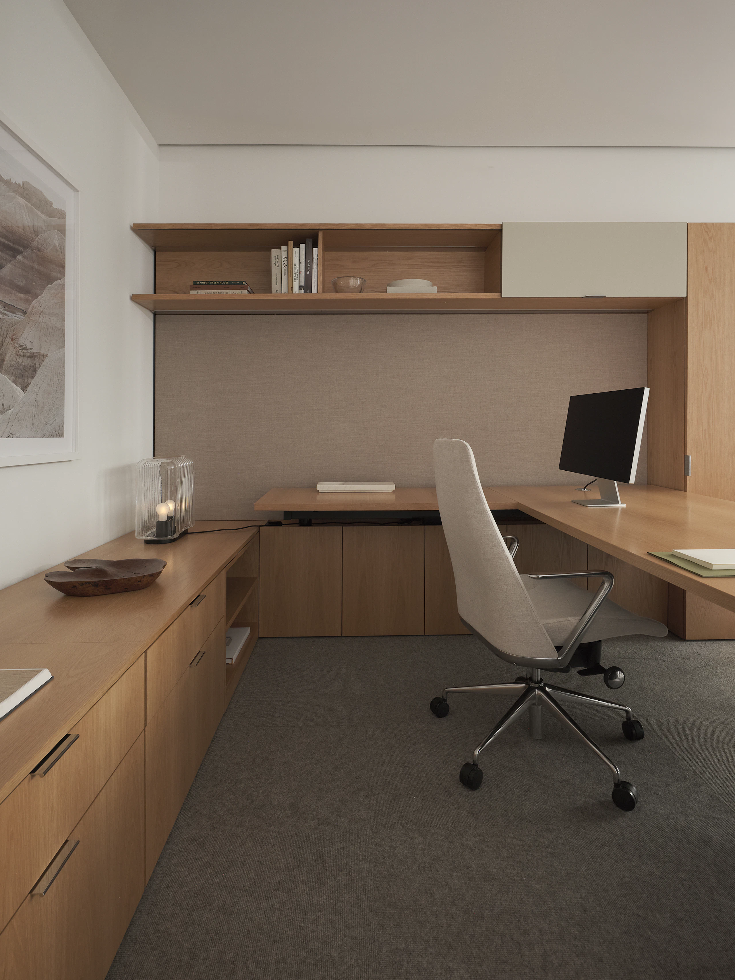 A Geiger One Private Office in Natural Flat Cut Oak with an L-shaped height adjustable work surface, credenza, overhead, and wardrobe storage, and a Taper Chair, viewed in profile.