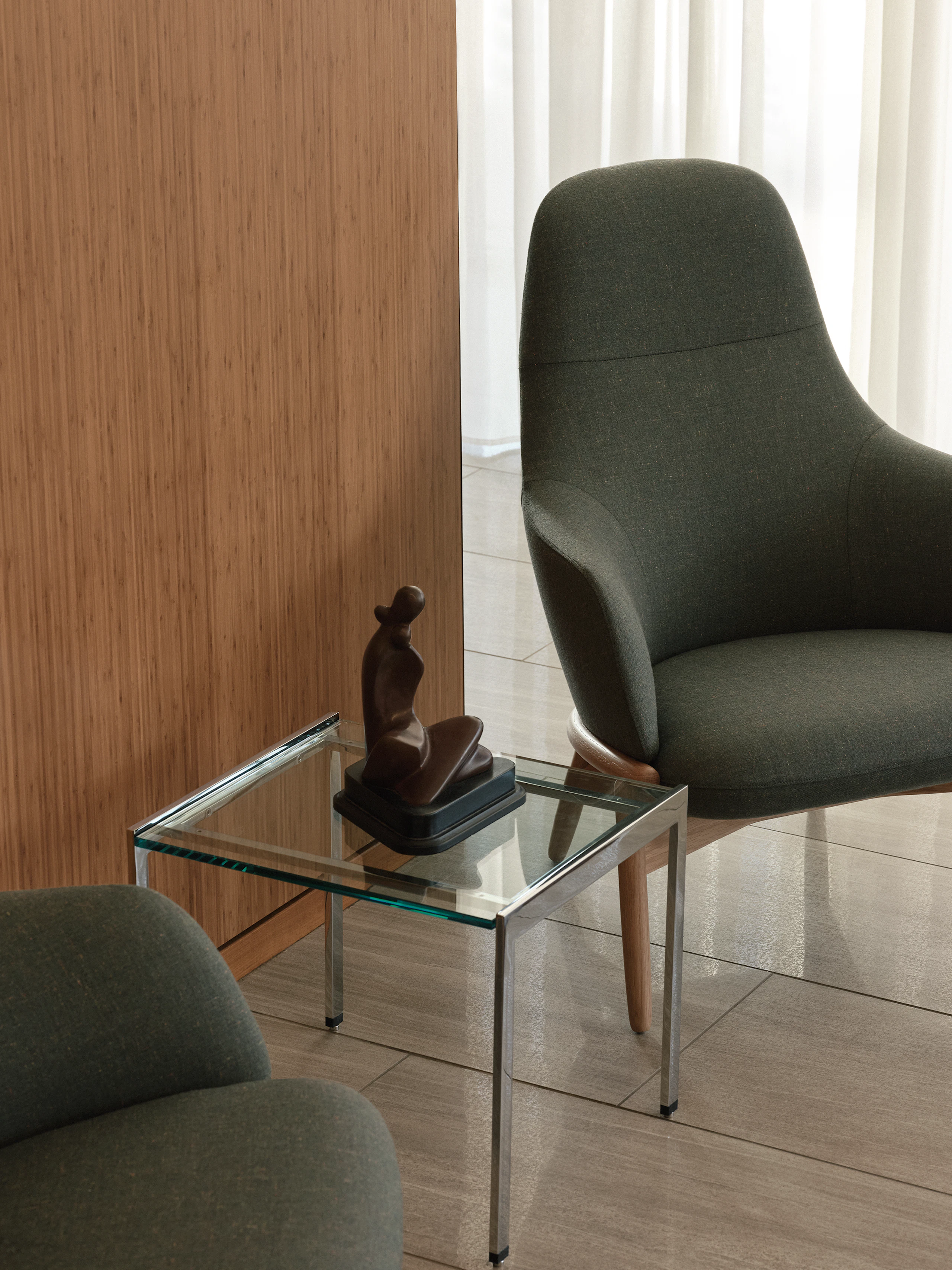 An H Frame Side Table with a clear glass top and chrome base between two high-back Reframe Lounge Chairs in Oak upholstered with Rowan in Zucchini, viewed at an angle against a wood wall.