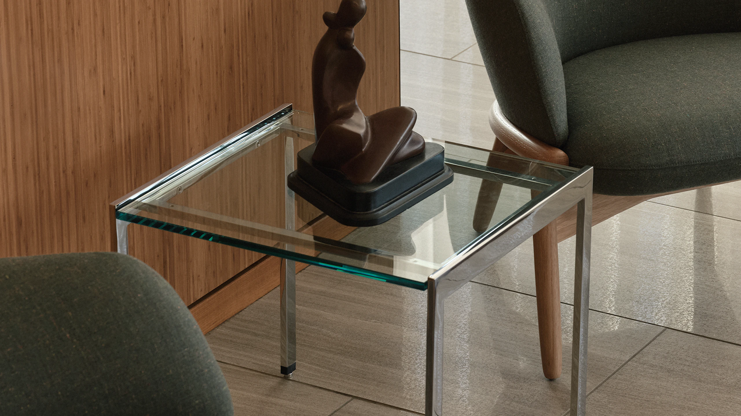 An H Frame Side Table with a clear glass top and chrome base between two high-back Reframe Lounge Chairs in Oak upholstered with Rowan in Zucchini, viewed at an angle against a wood wall.