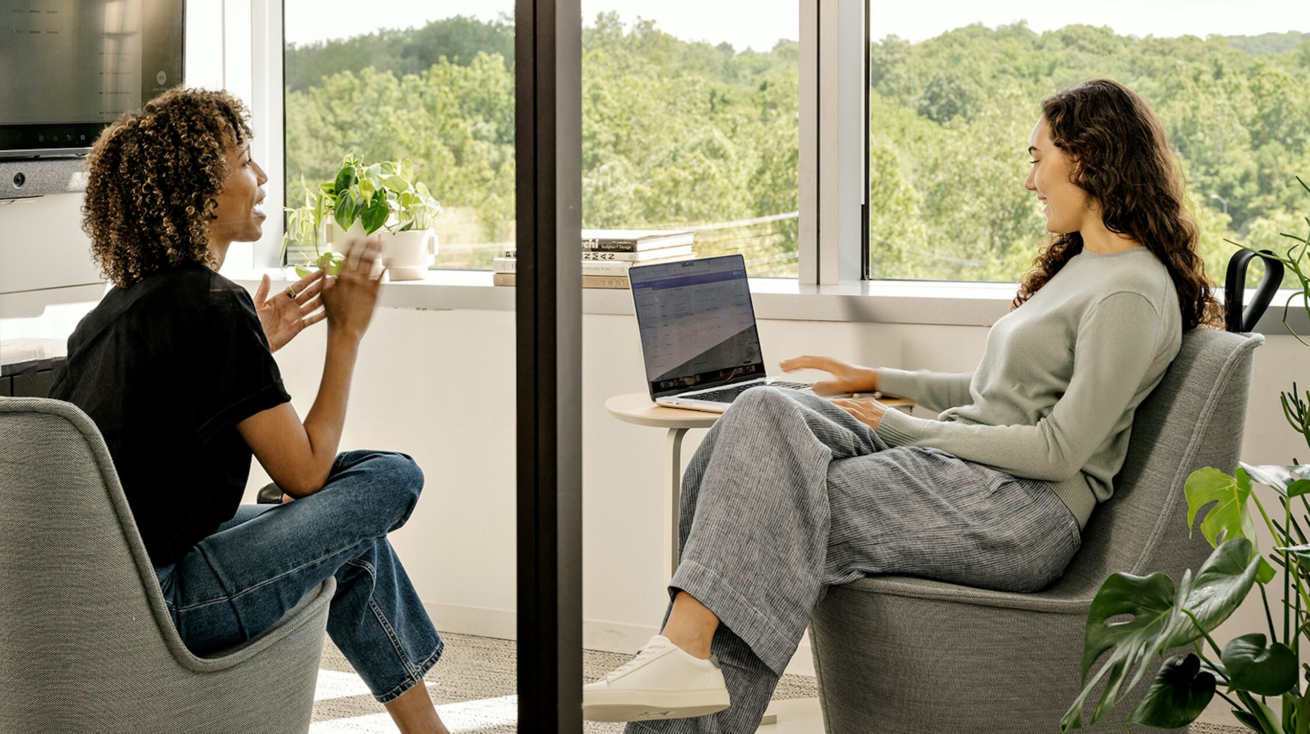 Two people, seated on grey chairs and overlooking green foliage outside large windows, having a cheerful conversation while one works on a laptop.