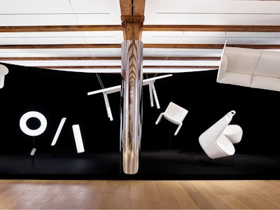A variety of white furniture suspended from the ceiling in front of a large black sheet at the NaughtOne showroom at MillerKnoll Design Days
