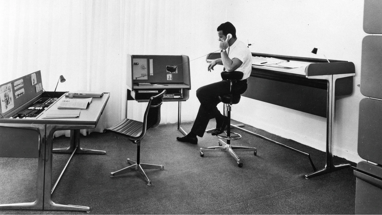 Black and white photo of a man talking on the phone while sitting in a counter-height stool at vintage desks.