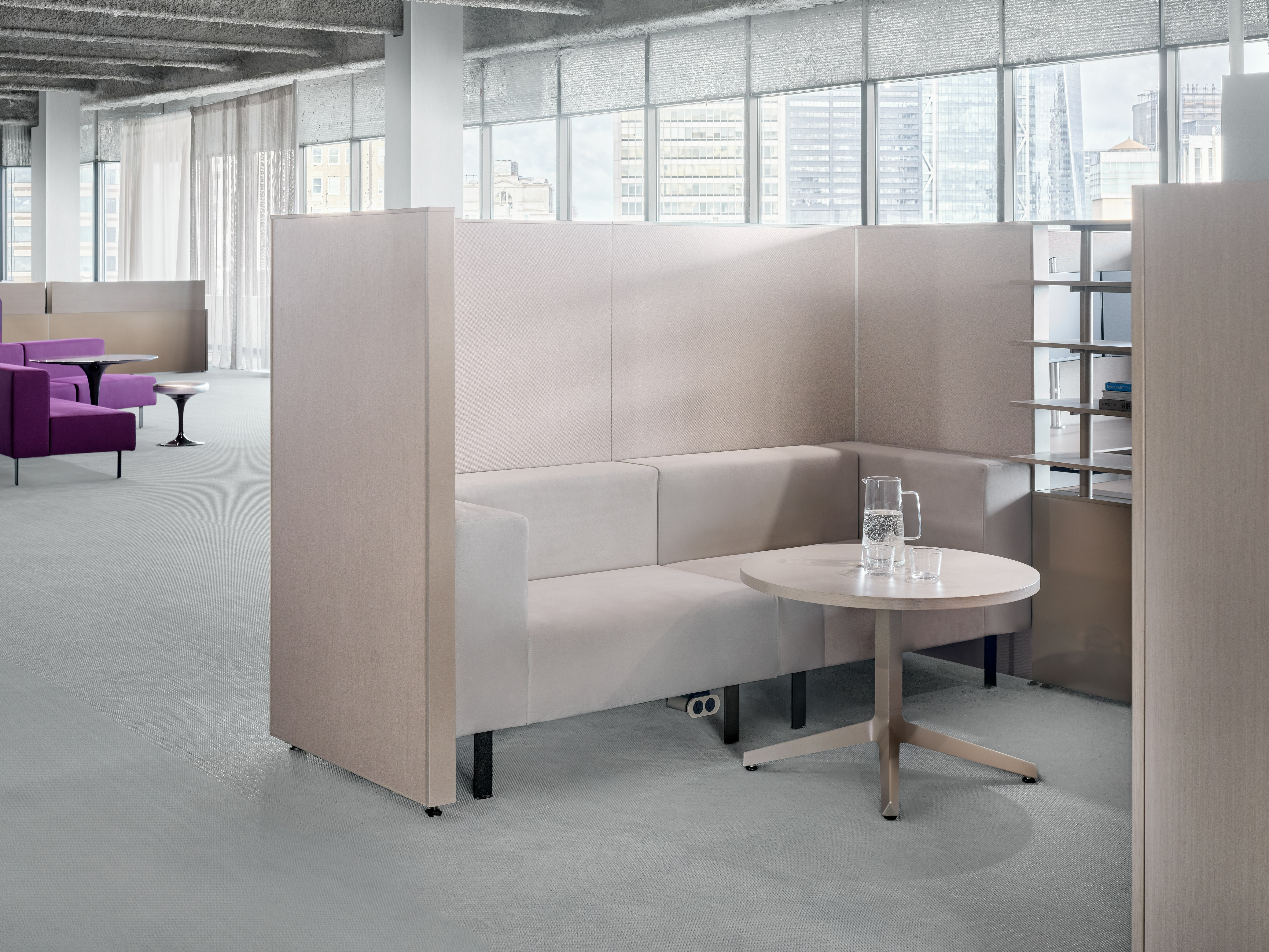 A working lounge setting is enclosed by privacy panels and has open shelves on one side. In the background there is an another open working lounge area.