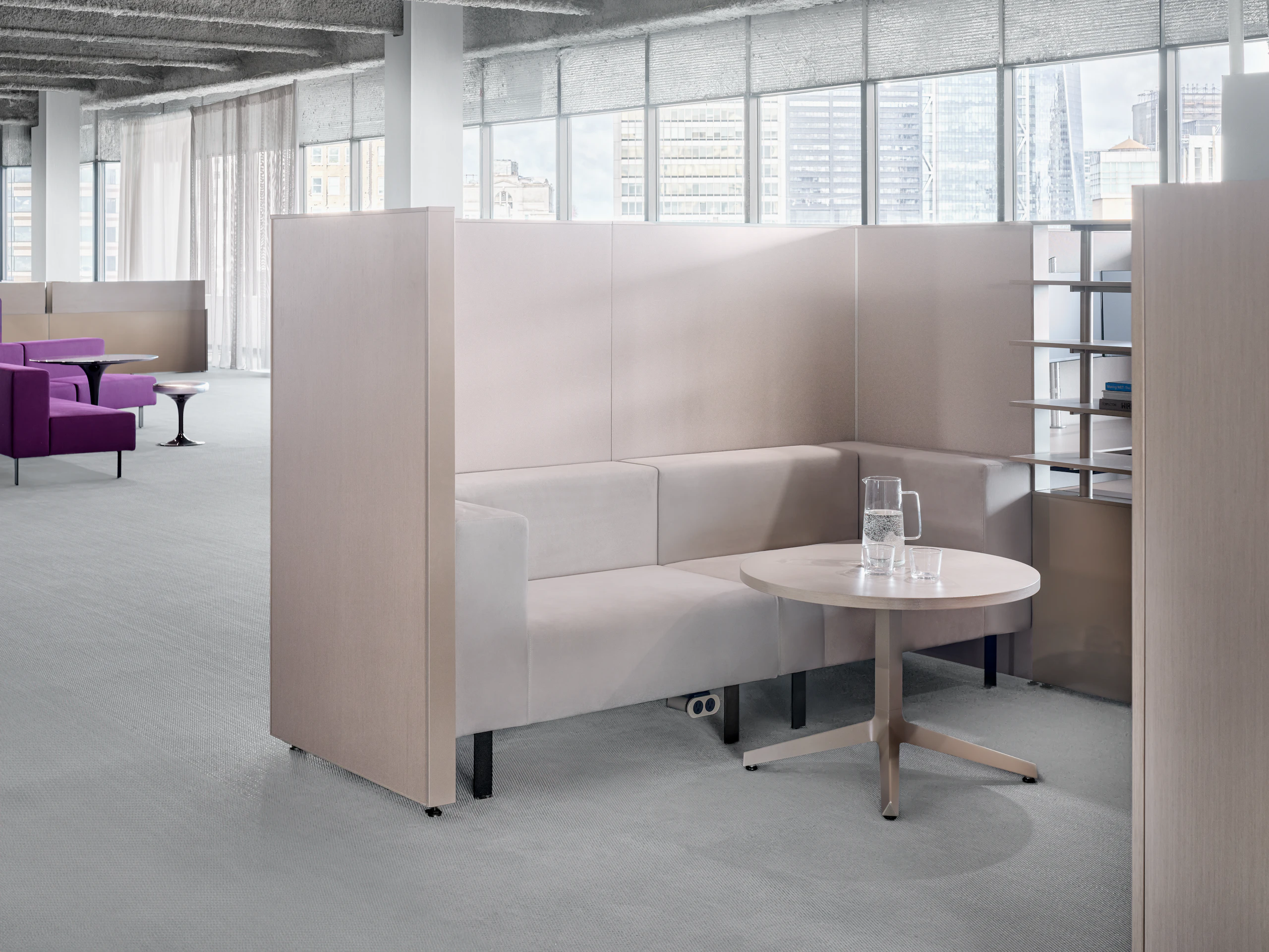 A working lounge setting is enclosed by privacy panels and has open shelves on one side. In the background there is an another open working lounge area.