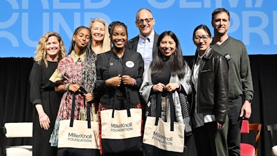 A group of people stand on a stage. Three young women hold black and white MillerKnoll Foundation tote bags.