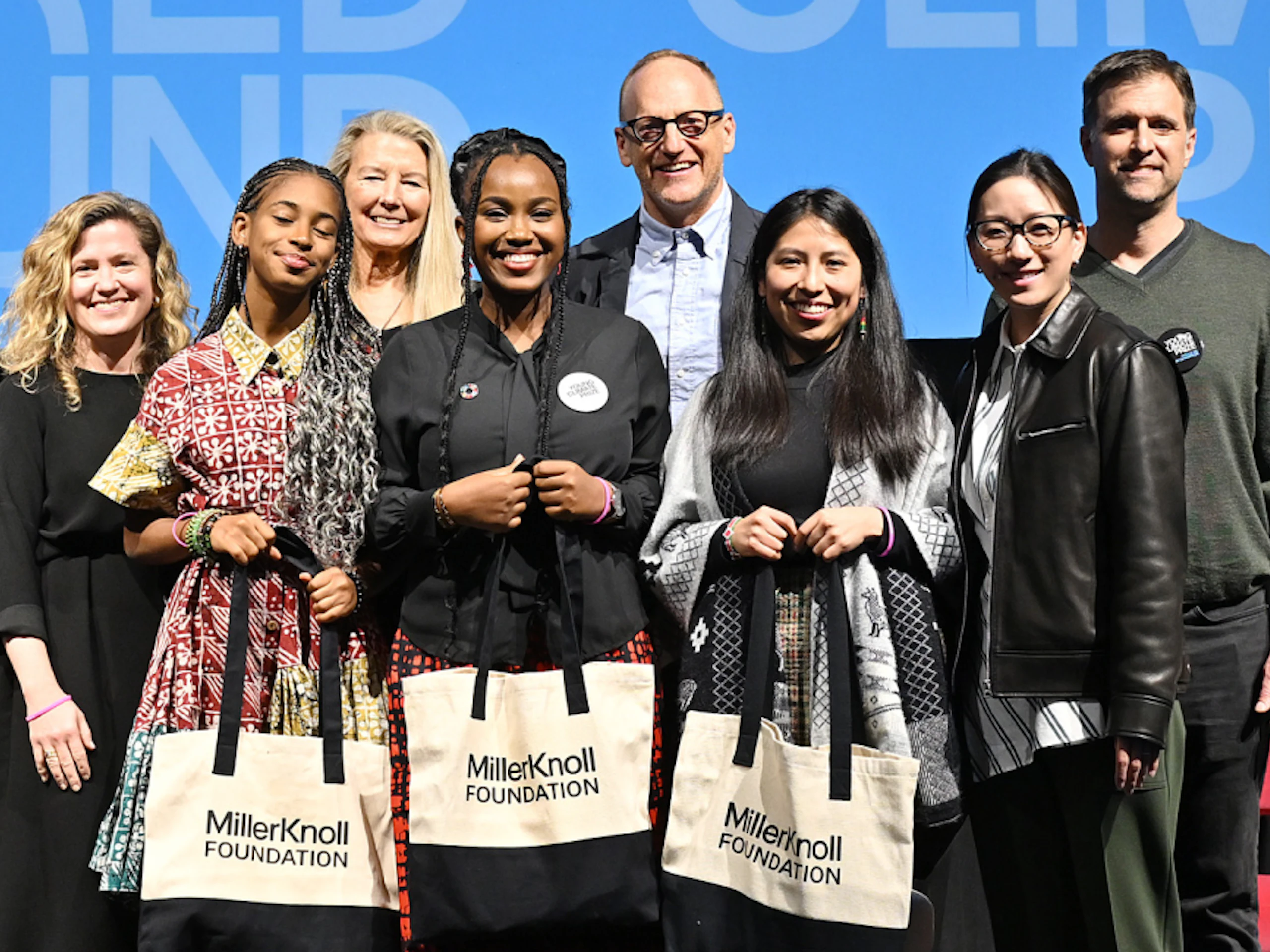 A group of people stand on a stage. Three young women hold black and white MillerKnoll Foundation tote bags.