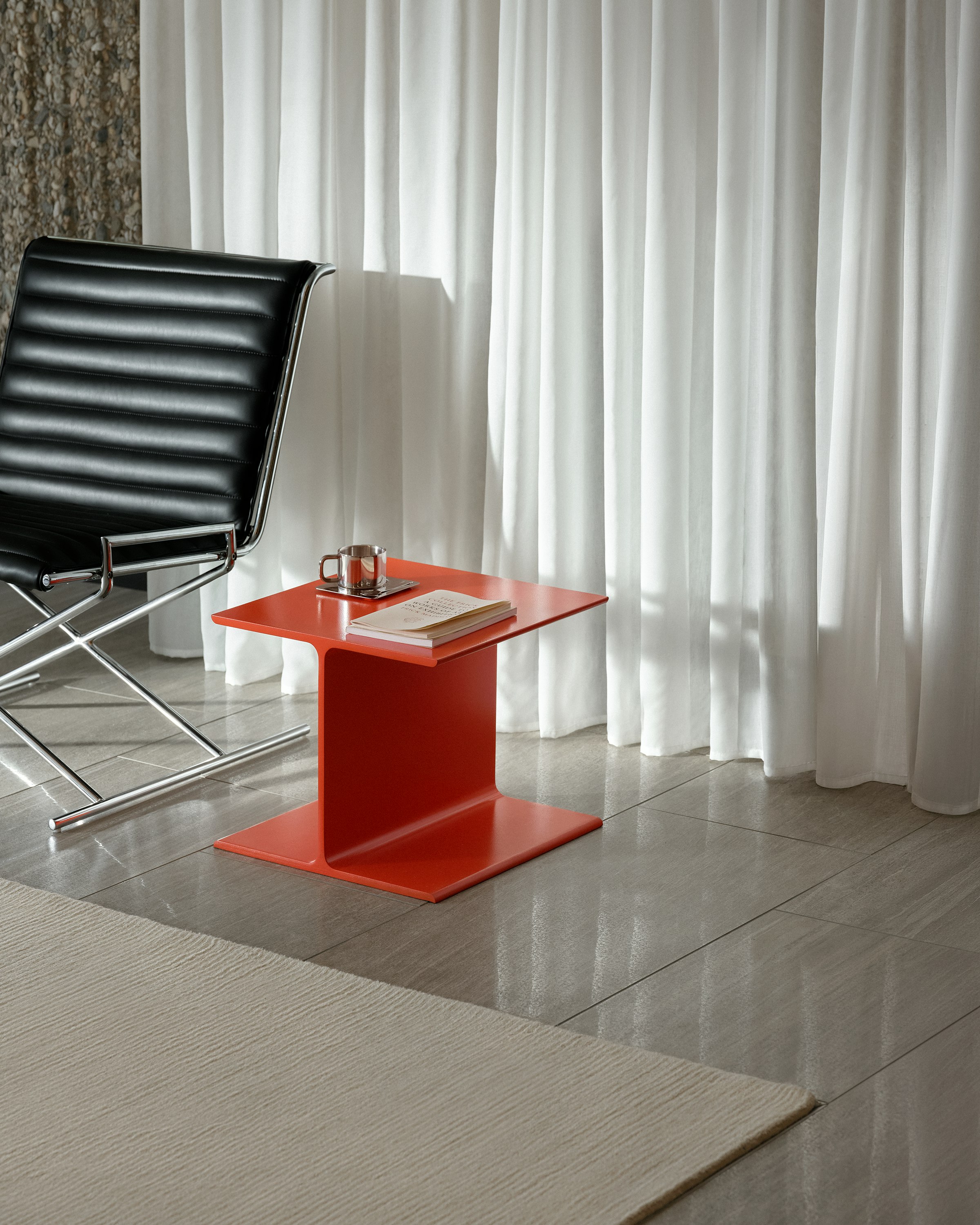 I Beam Side Table in International Orange beside a Sled Chair upholstered with black leather with a chrome frame, viewed at an angle against a white curtain and stone wall.