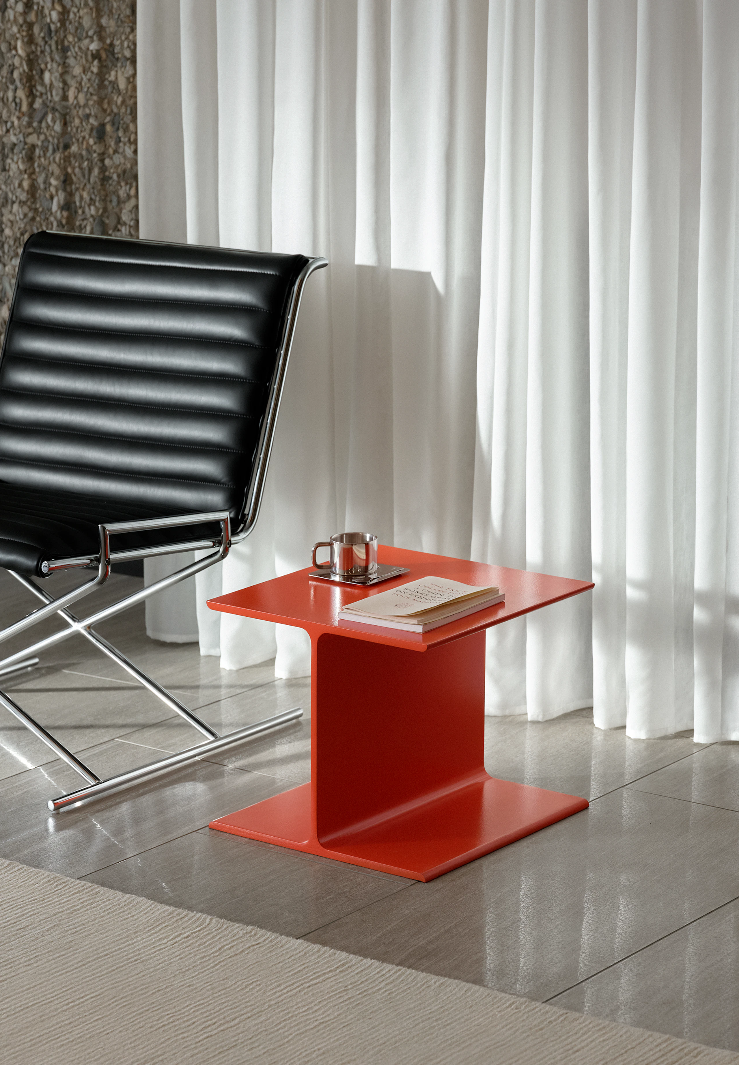 I Beam Side Table in International Orange beside a Sled Chair upholstered with black leather with a chrome frame, viewed at an angle against a white curtain and stone wall.