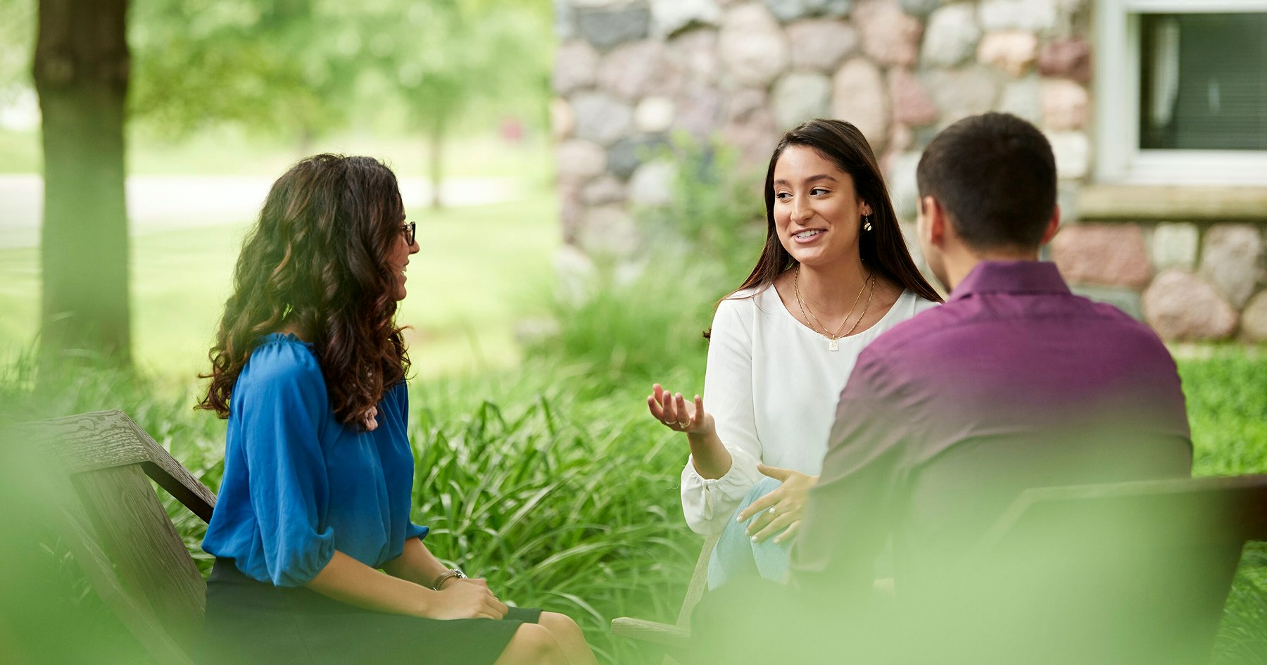 Three people seated in wooden chairs having a conversation outside next to some green foliage and a stone house.