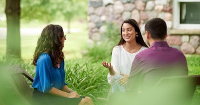 Three people seated in wooden chairs having a conversation outside next to some green foliage and a stone house.