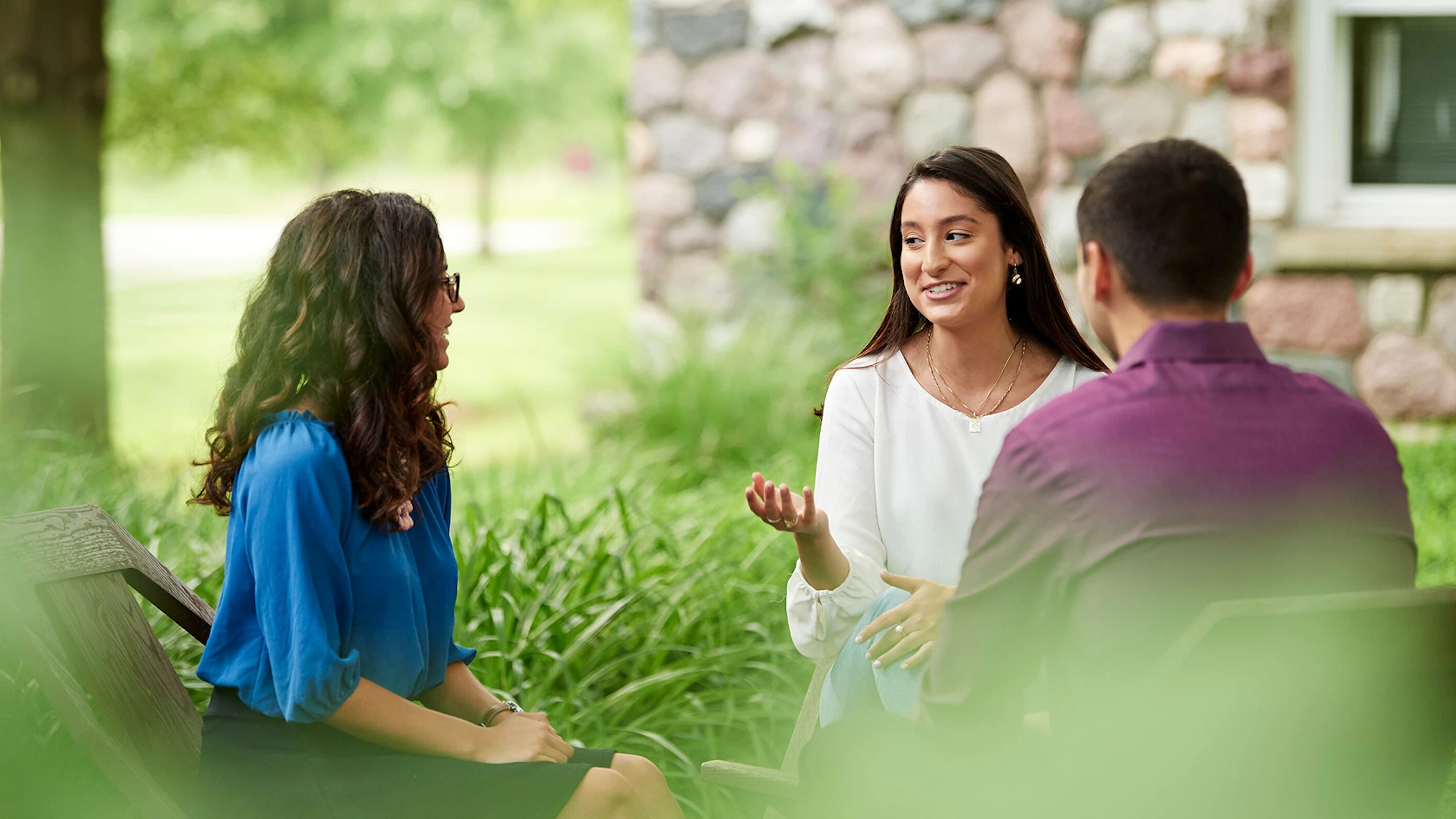 Three people seated in wooden chairs having a conversation outside next to some green foliage and a stone house.