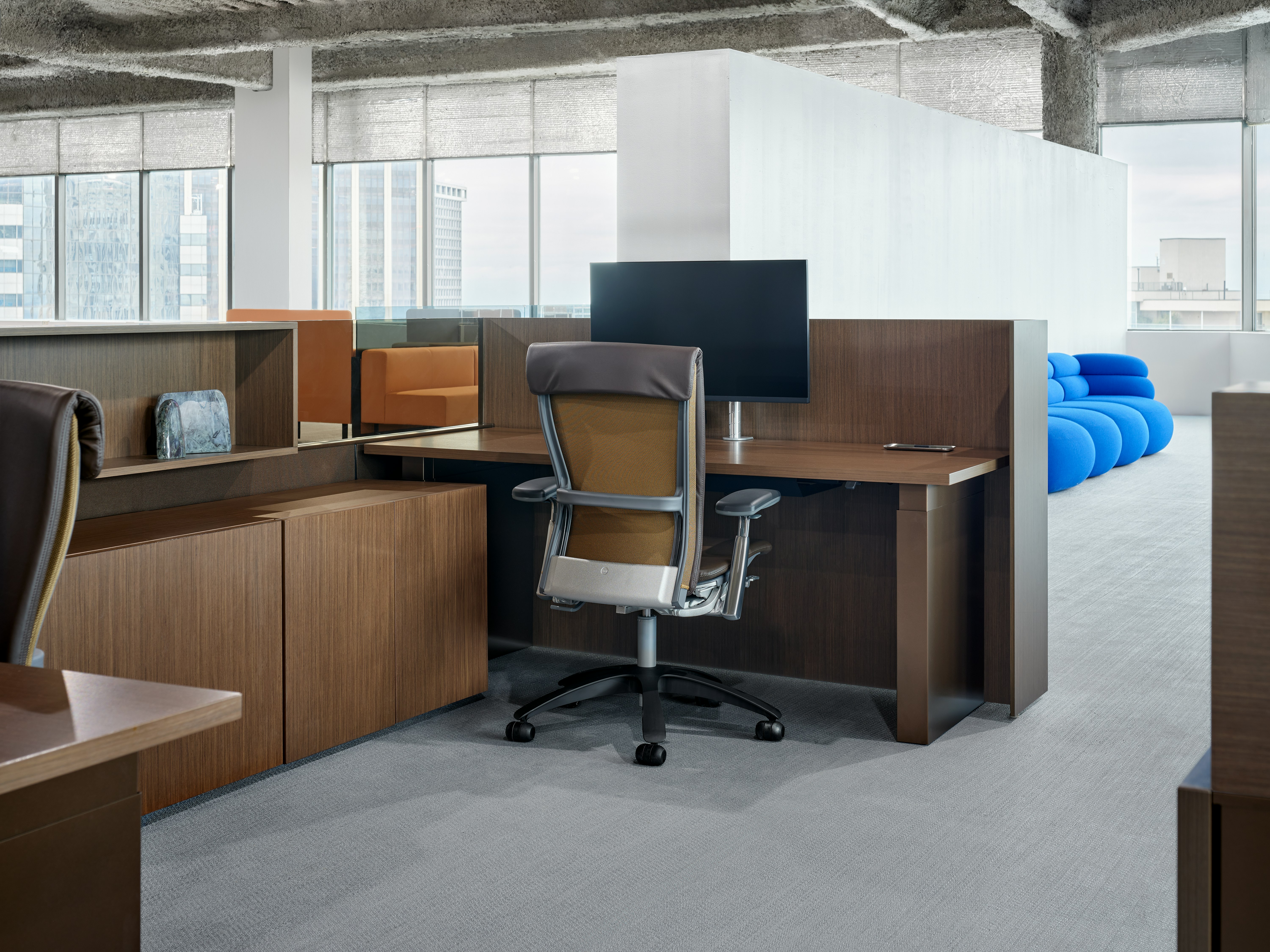 A work chair is sitting in front of a height adjustable table, with some storage cubbies and credenzas next to the work surface. In the background, there is some lounge furniture.