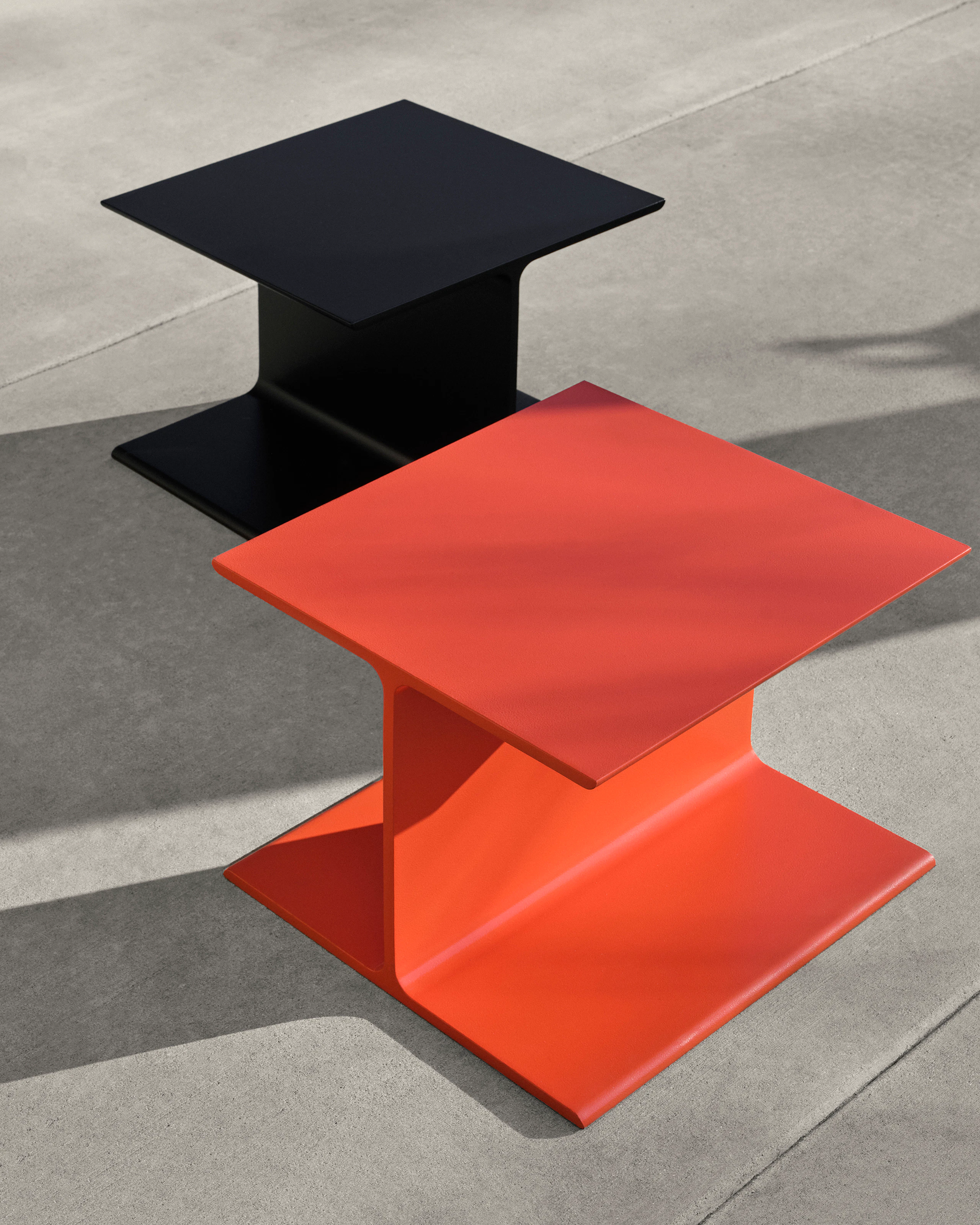 Two I Beam Side Tables designed by Ward Bennett, one in International Orange positioned slightly in front of one in black, viewed from above at an angle on a concrete floor.