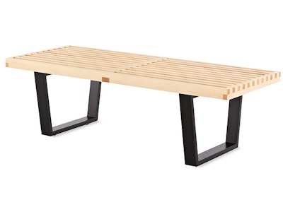 A front three quarter view of a natural maple Nelson Platform Bench by Herman Miller with black metal legs on a white background