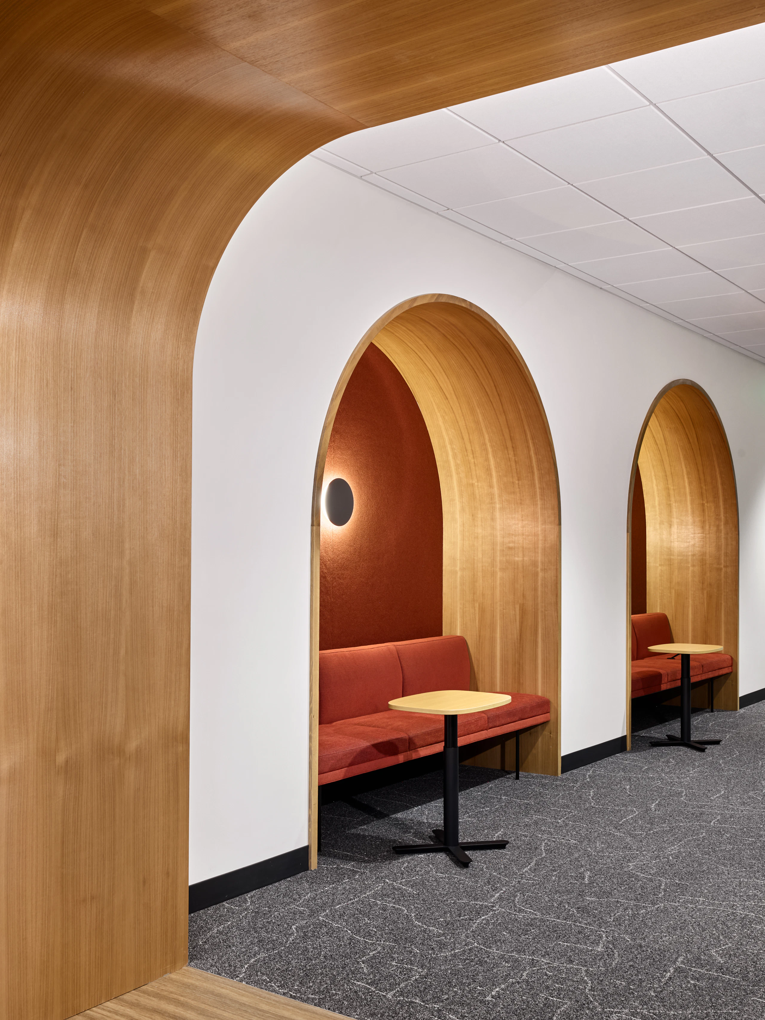 Wood-accented corridor with arched upholstered alcoves, integrated lounge seating, and Herman Miller Passport Tables.
