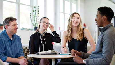 Four people seated on a couch around a small round table. Two are laughing while another is speaking.
