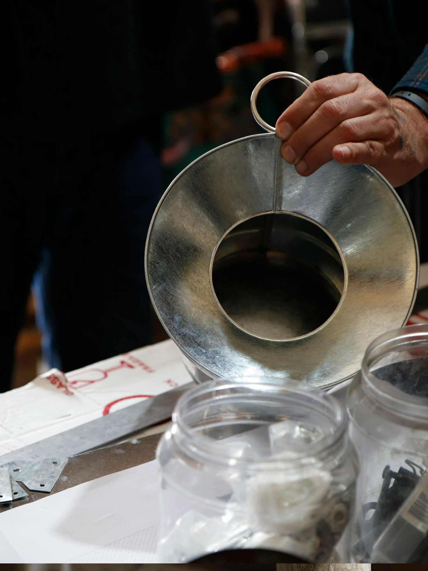 A person crafting a bare metal watering can at a workbench. Various parts and materials, some in glass jars, can be seen across the table.