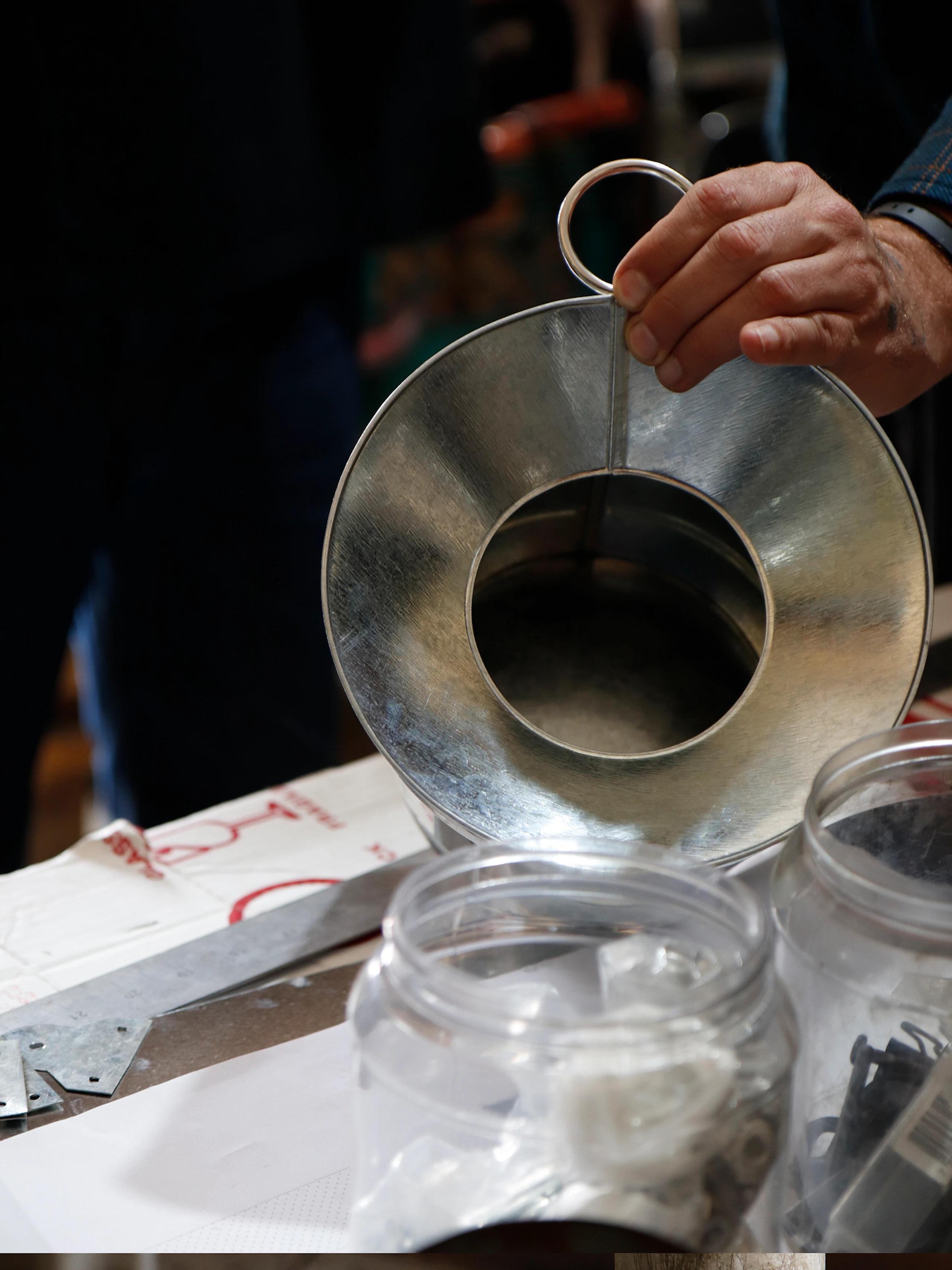A person crafting a bare metal watering can at a workbench. Various parts and materials, some in glass jars, can be seen across the table.
