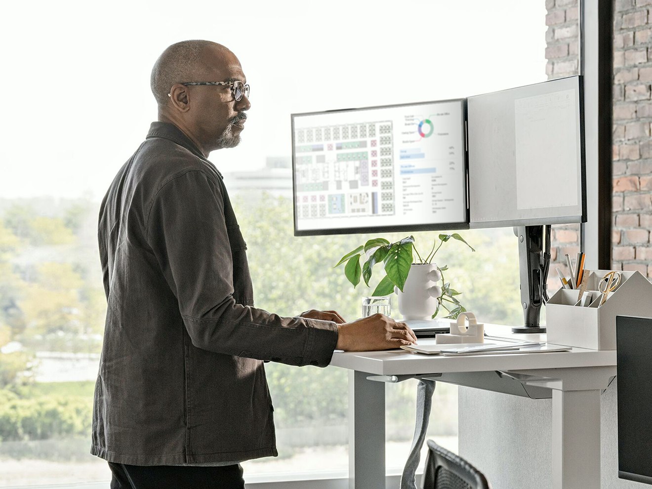 Man standing at a sit-to-stand desk while working on two computer monitors.