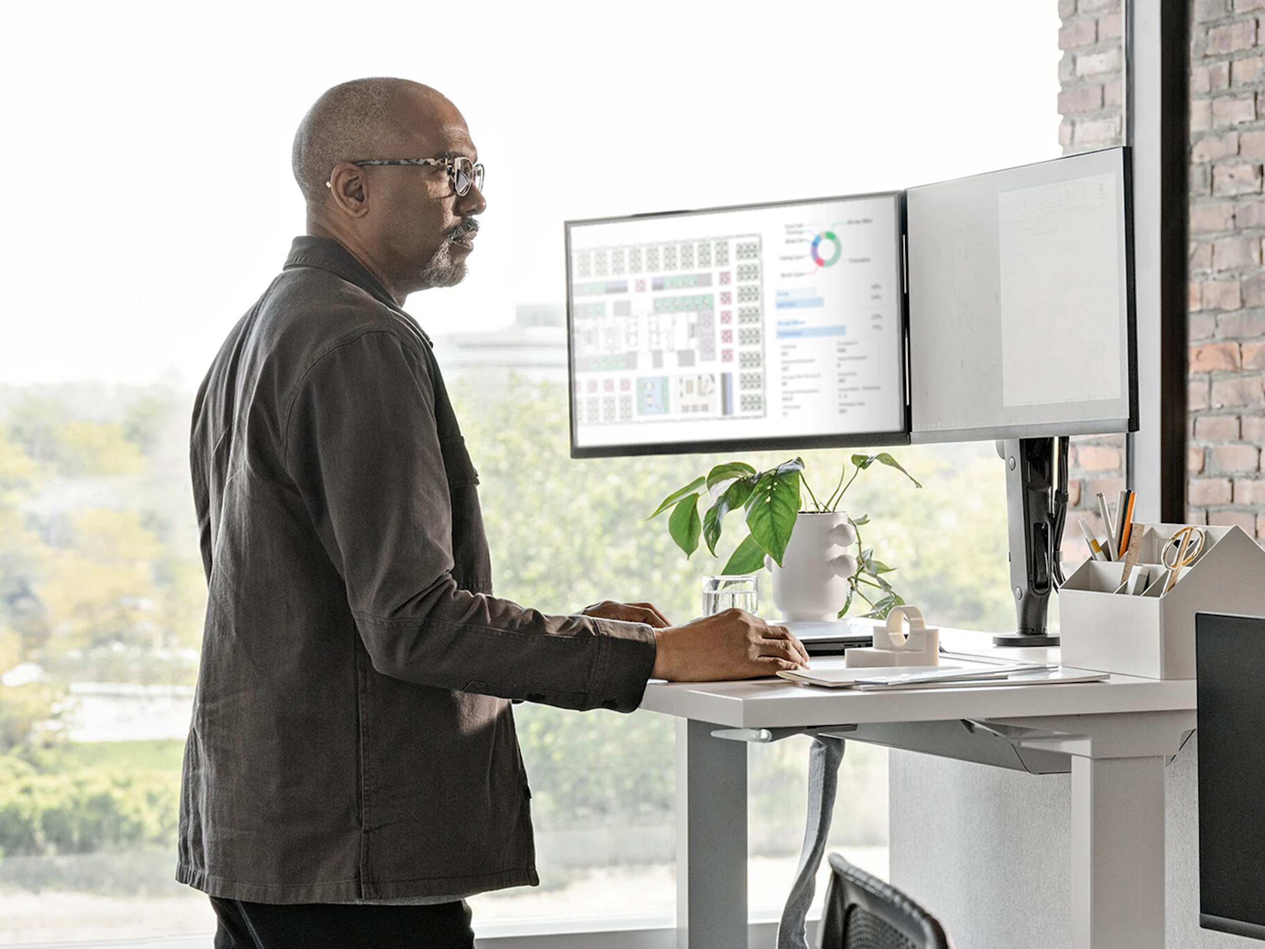 Man standing at a sit-to-stand desk while working on two computer monitors.