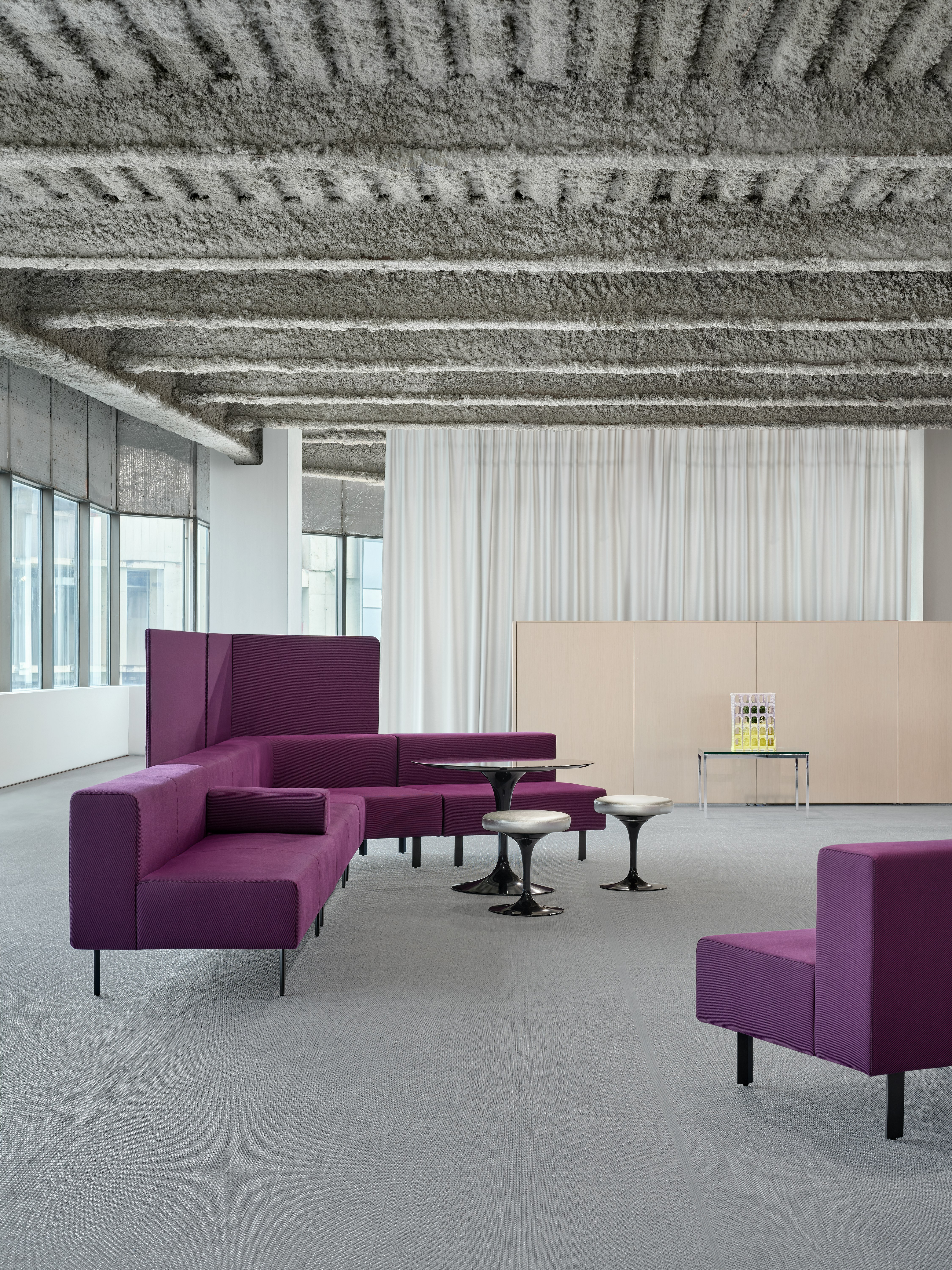 A bright purple, working lounge setting is positioned in an open office. There are also a table and stools in front of the lounge bench.
