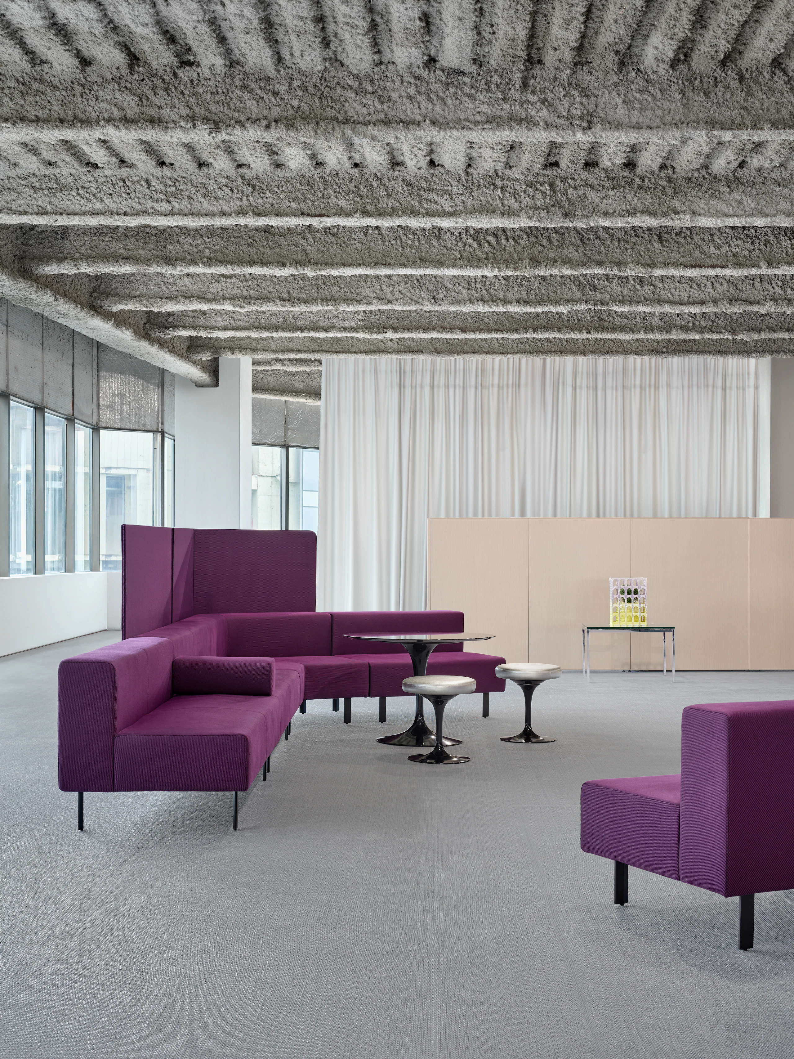 A bright purple, working lounge setting is positioned in an open office. There are also a table and stools in front of the lounge bench.