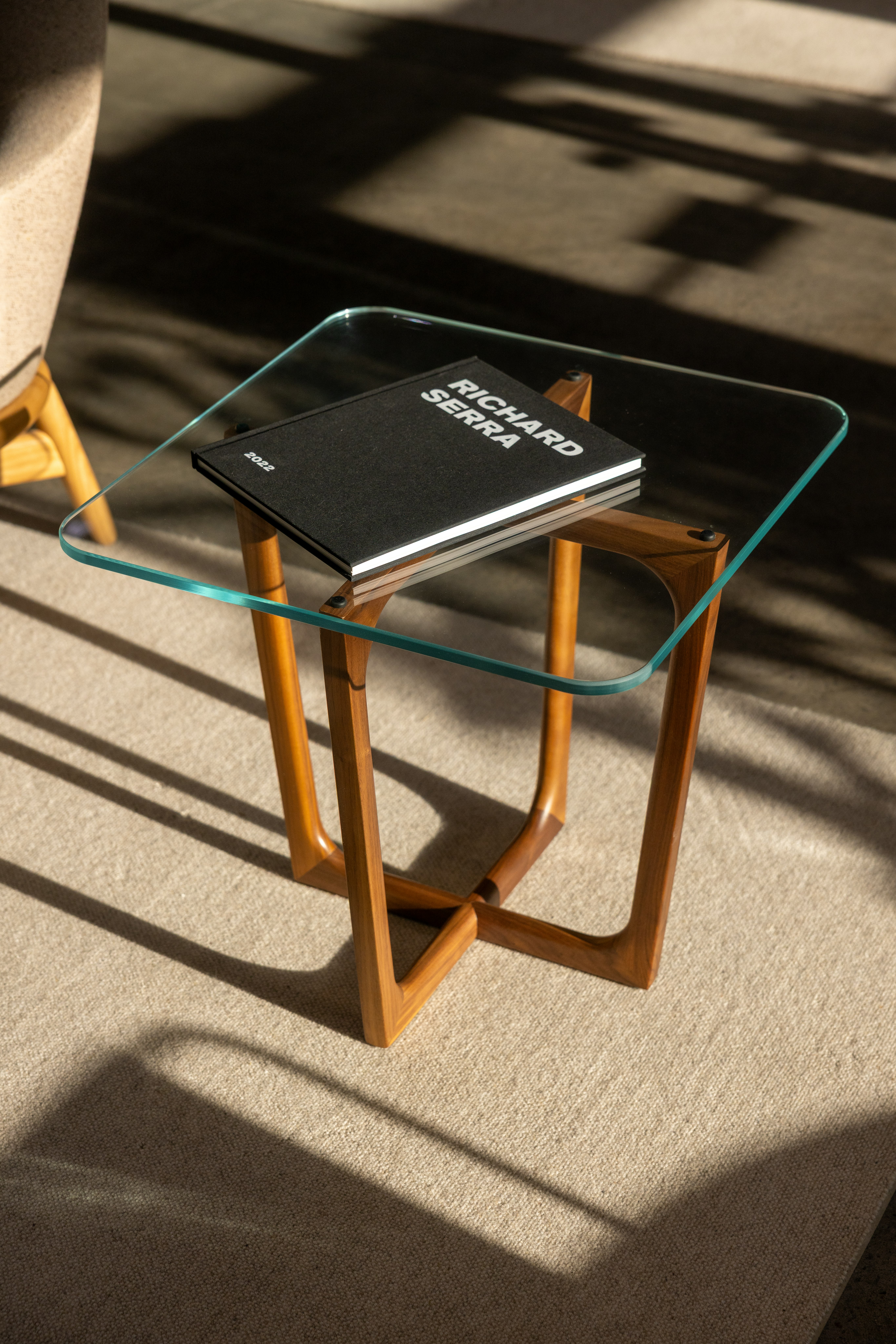 Loophole Side Table with a square clear glass top and Walnut base, styled with a black book on top, viewed from above at an angle in a showroom setting.