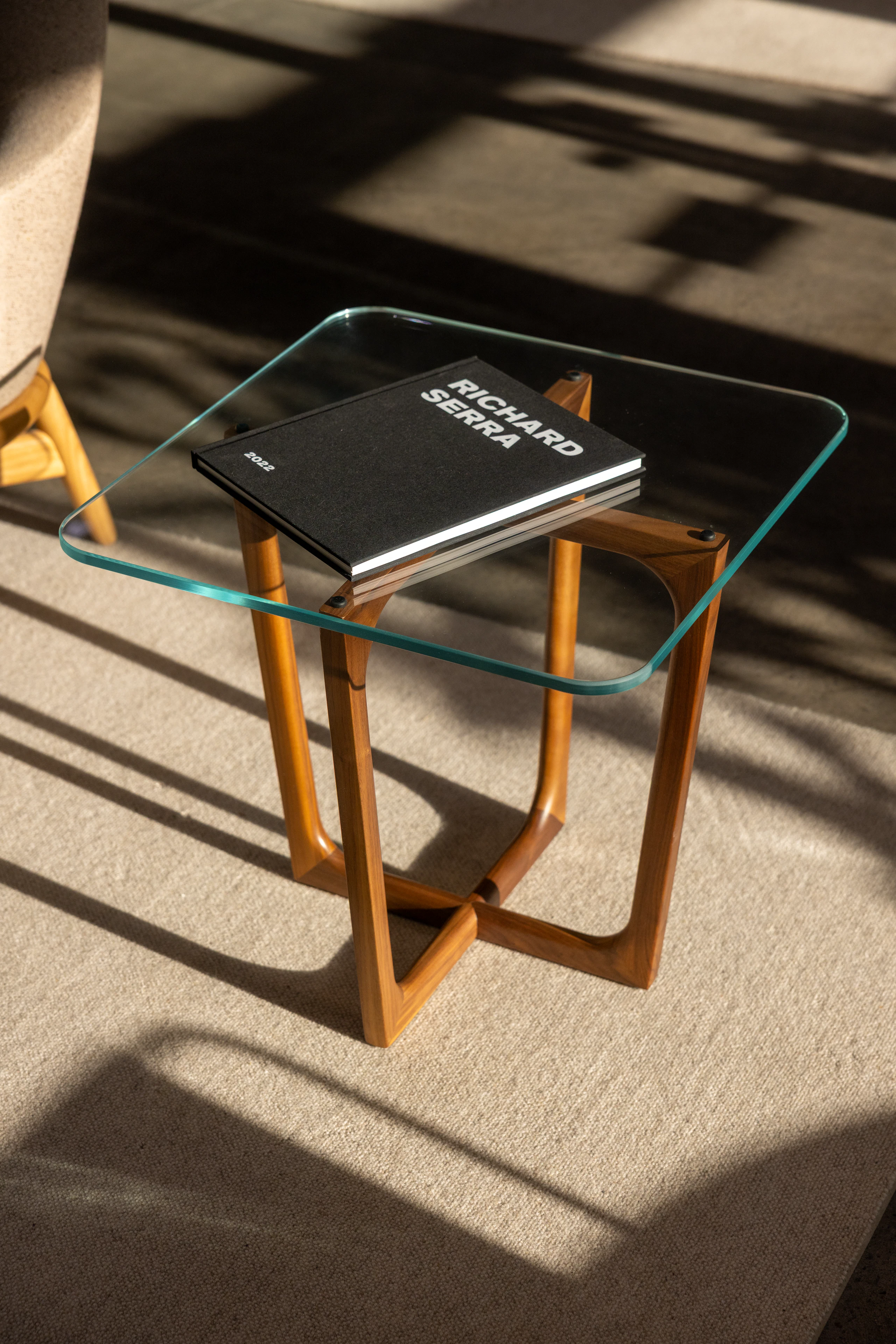 Loophole Side Table with a square clear glass top and Walnut base, styled with a black book on top, viewed from above at an angle in a showroom setting.