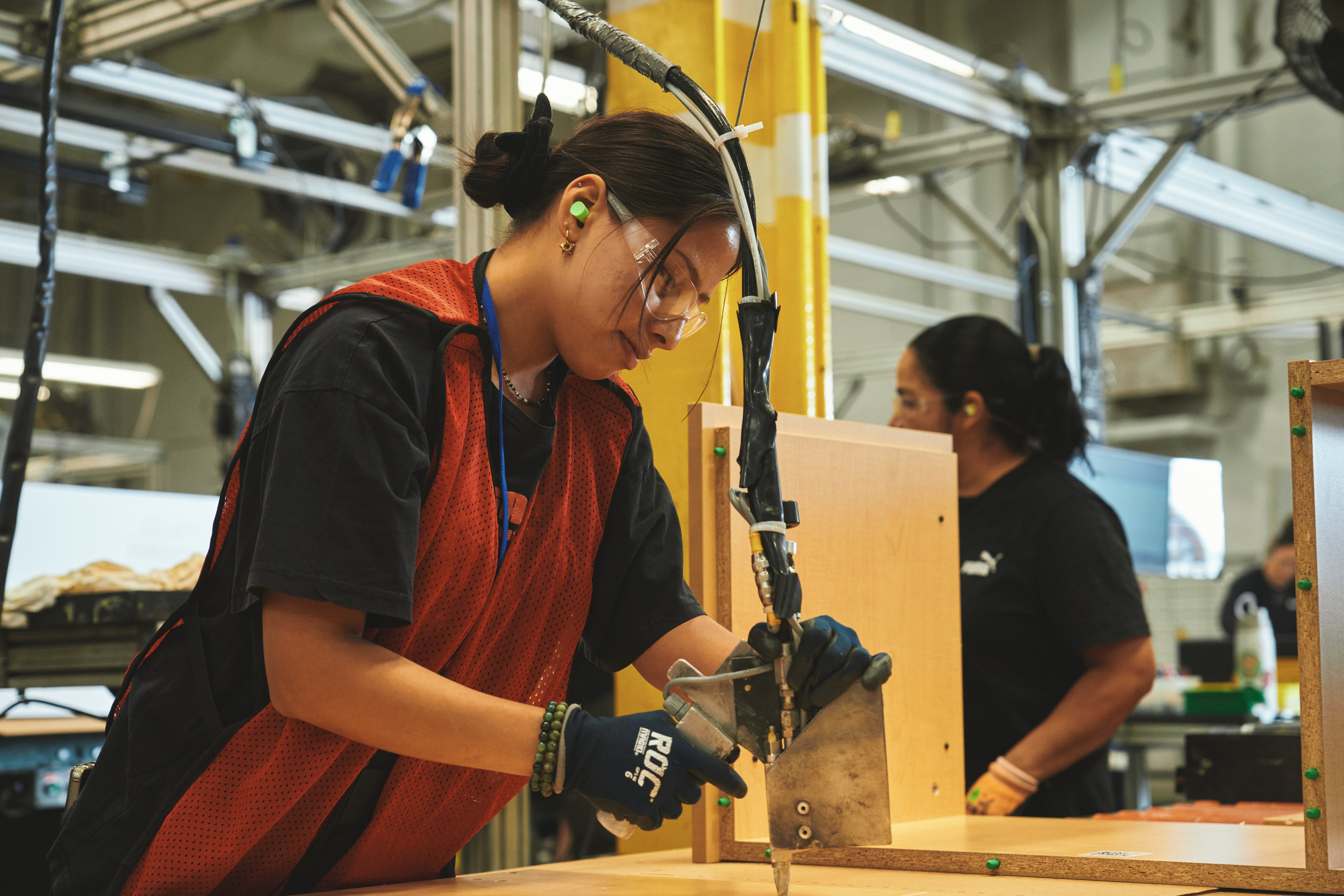A woman wearing safety glasses, a red vest, a black shirt, and black gloves works on a furniture assembly line. Pieces of wood with green dowels can be seen at her station.