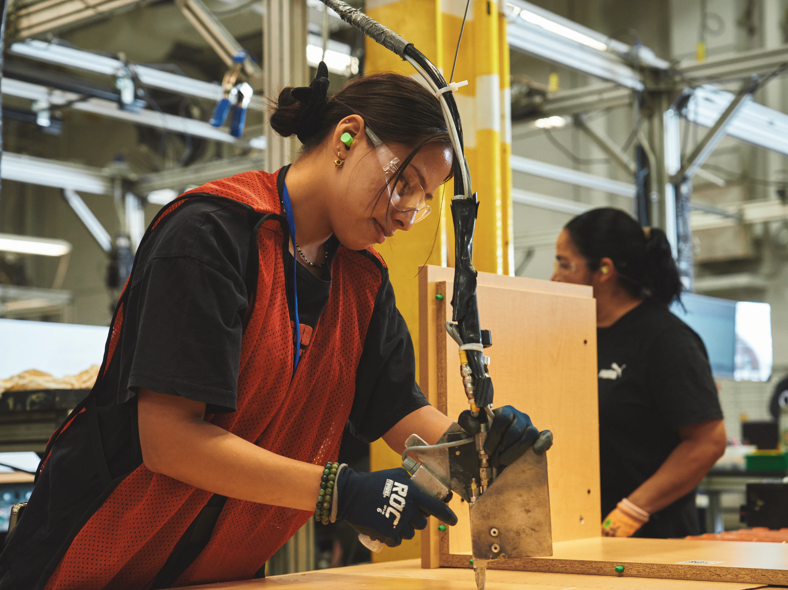 A woman wearing safety glasses, a red vest, a black shirt, and black gloves works on a furniture assembly line. Pieces of wood with green dowels can be seen at her station.