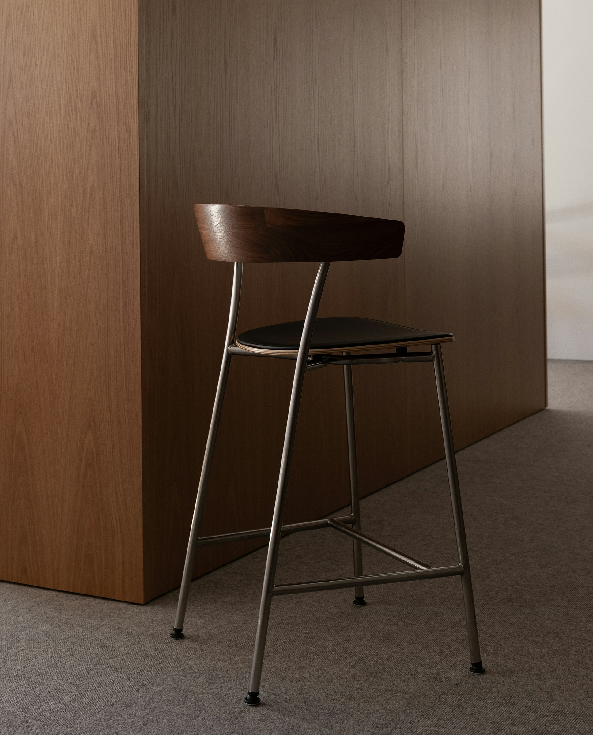 Counter height Leeway Stool designed by Keiji Takeuchi in walnut with a satin chrome frame and black leather seat, viewed from behind at an angle in an office setting.