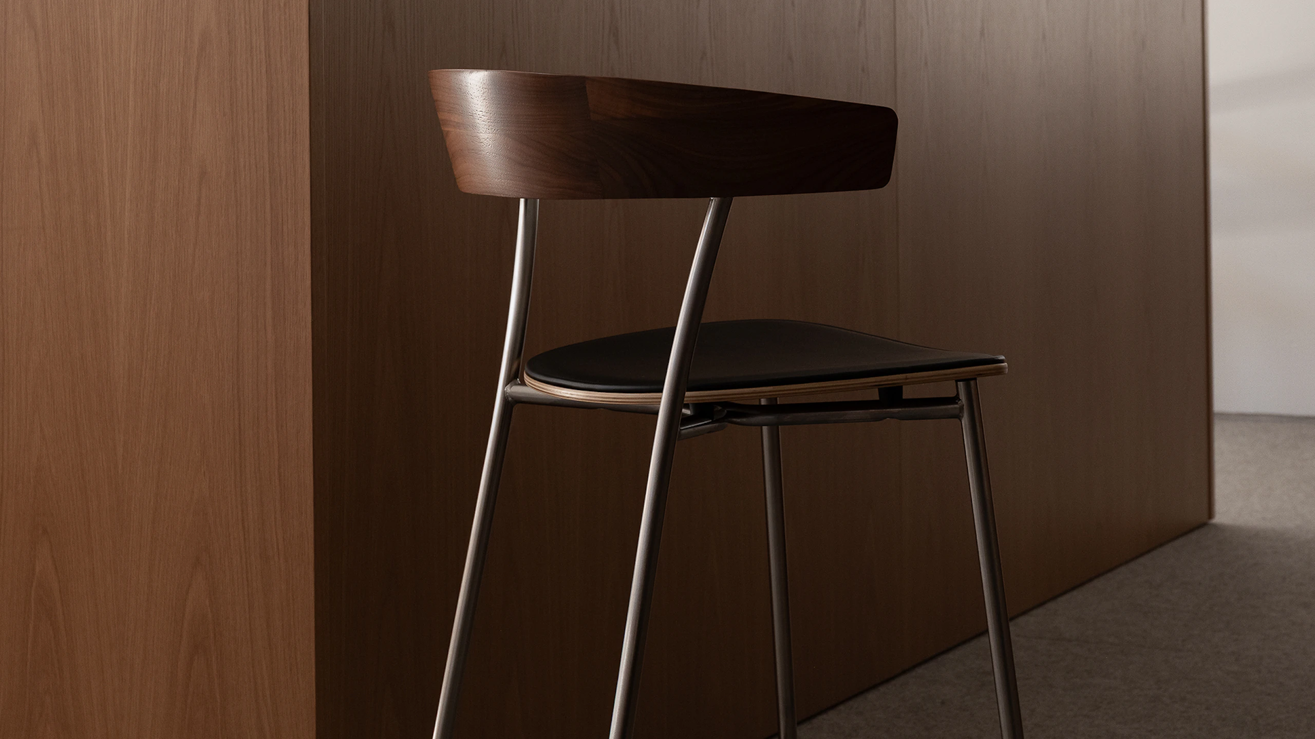 Counter height Leeway Stool designed by Keiji Takeuchi in walnut with a satin chrome frame and black leather seat, viewed from behind at an angle in an office setting.