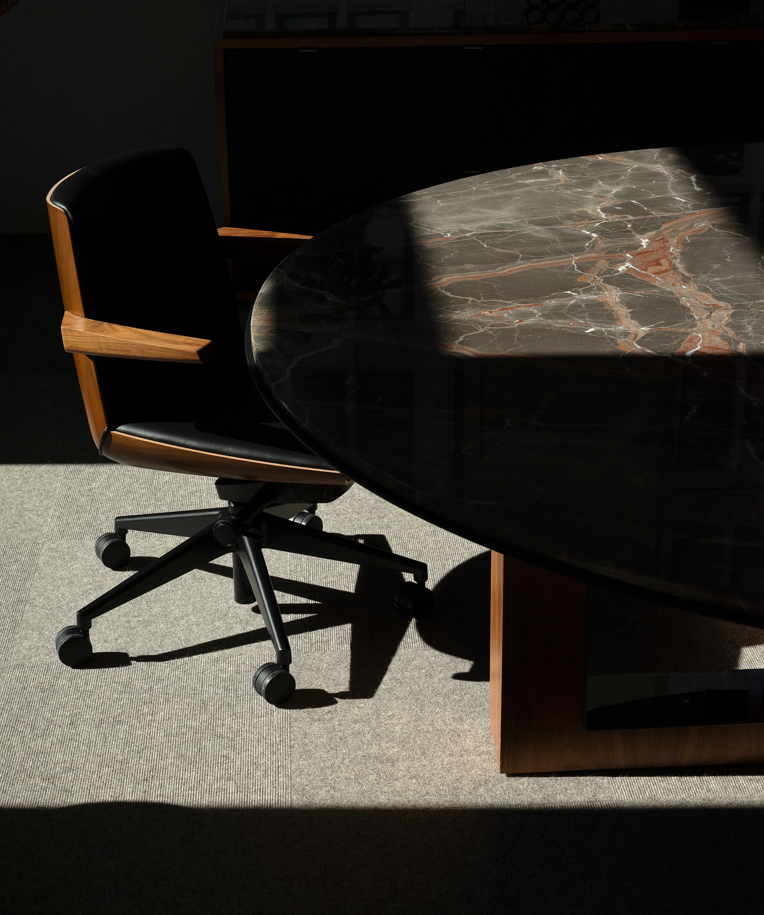 A low back Clamshell Conference Chair in Walnut, upholstered with black leather, beside a Highline Fifty Meeting Table with an Obra di Caravaggio Marble tabletop.