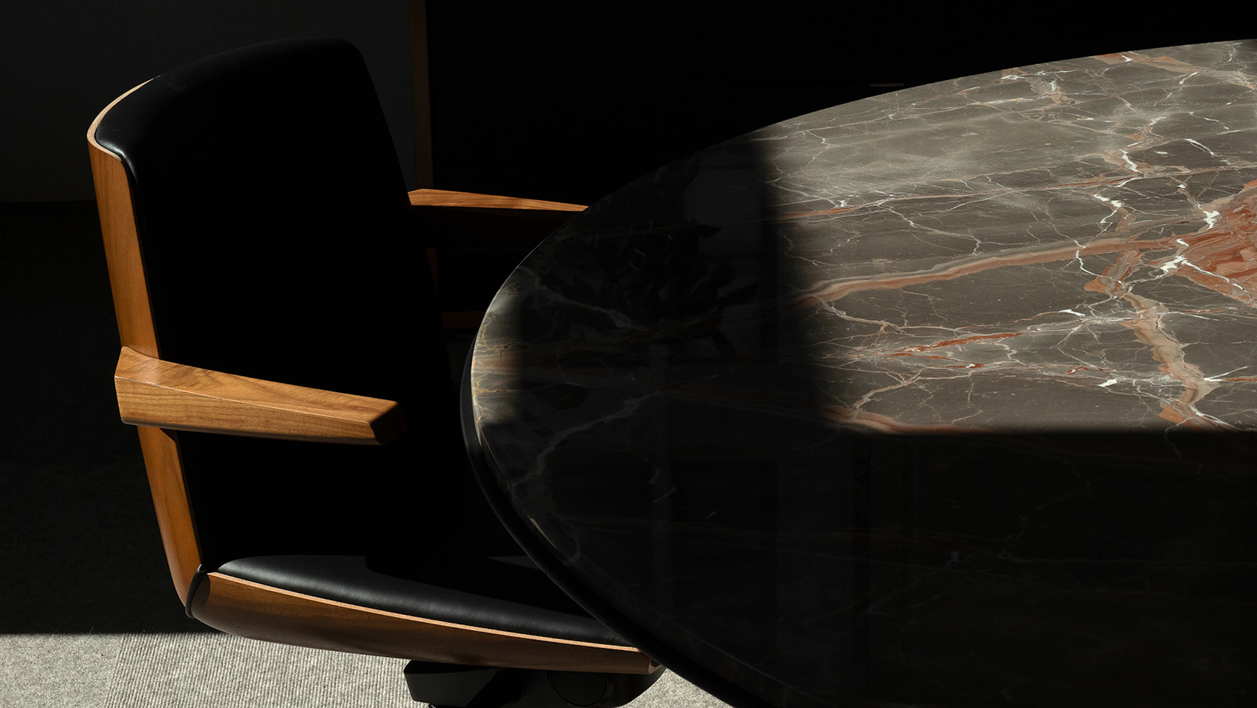 A low back Clamshell Conference Chair in Walnut, upholstered with black leather, beside a Highline Fifty Meeting Table with an Obra di Caravaggio Marble tabletop.