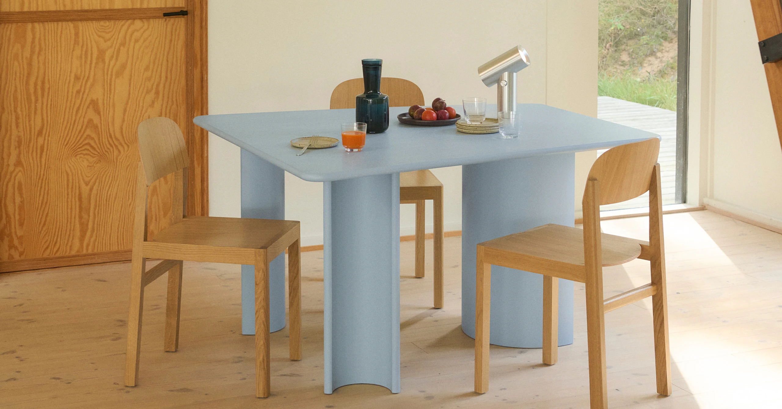 Light blue Re-norm table, Oak Workshop Chair, Polished Aluminum Beam Portable Lamp, and Dark Blue Raise Carafe in a warmly lit dining setting.