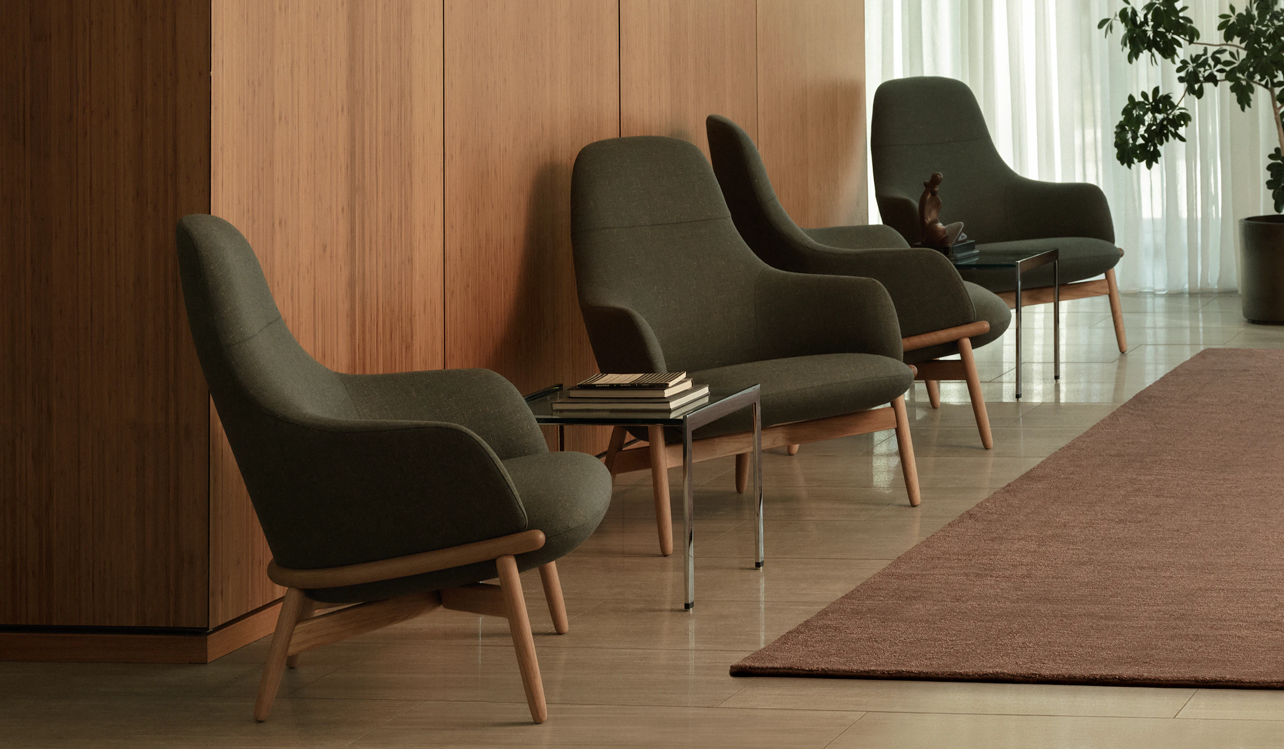 Four high-back Reframe Lounge Chairs in Oak upholstered with Rowan in Zucchini with two H Frame Side Tables, viewed at an angle against a wood wall.