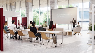 Personas sentadas en un aula moderna viendo una presentación en una pantalla grande. Algunos tienen computadoras portátiles abiertos sobre las mesas.