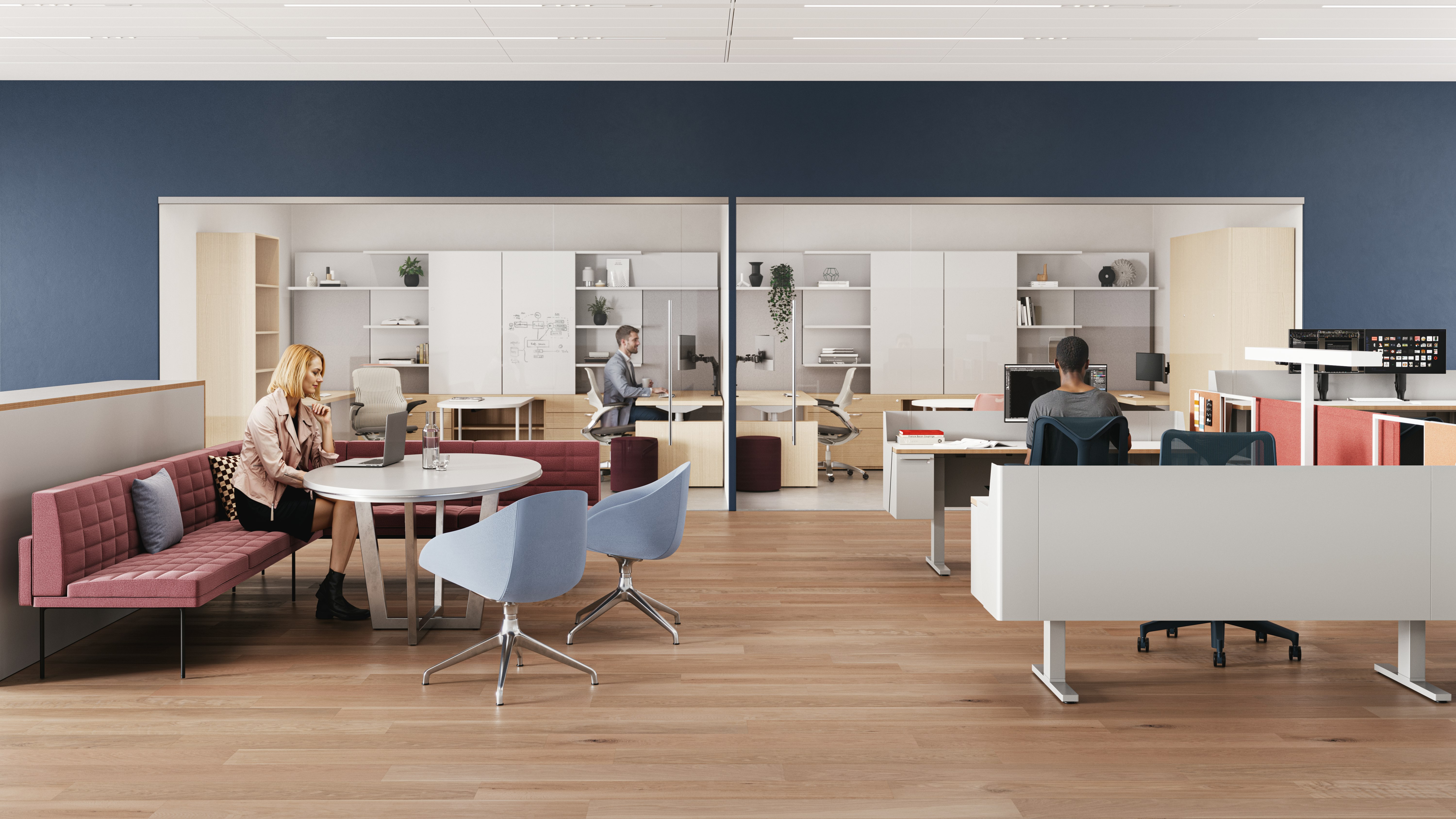  A modern workspace with wooden flooring, featuring an individual seated at a round table, along with a person working at a desk behind glass, and contemporary furniture, including red and blue chairs.
