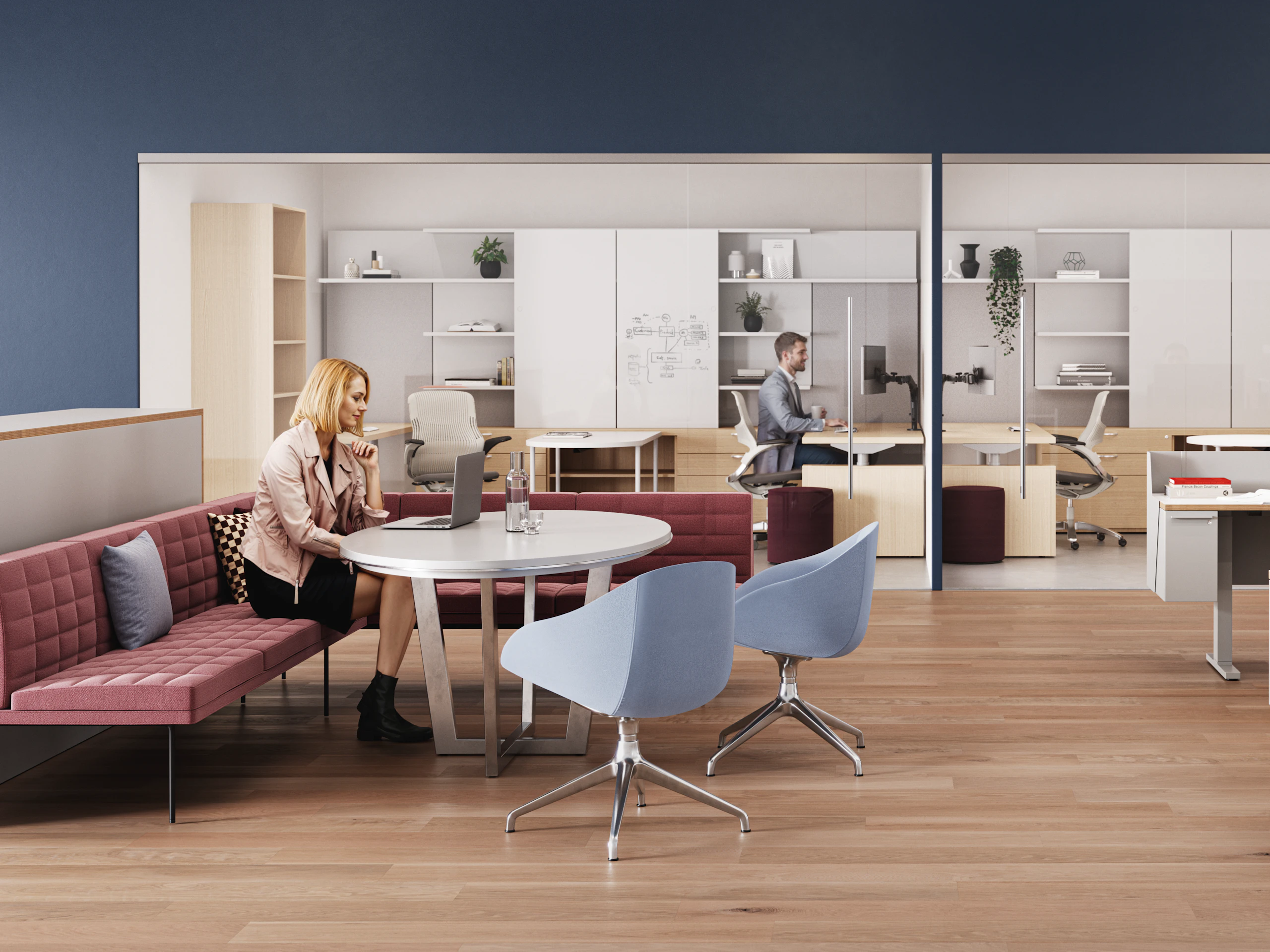 A modern workspace with wooden flooring, featuring an individual seated at a round table, along with a person working at a desk behind glass, and contemporary furniture, including red and blue chairs.