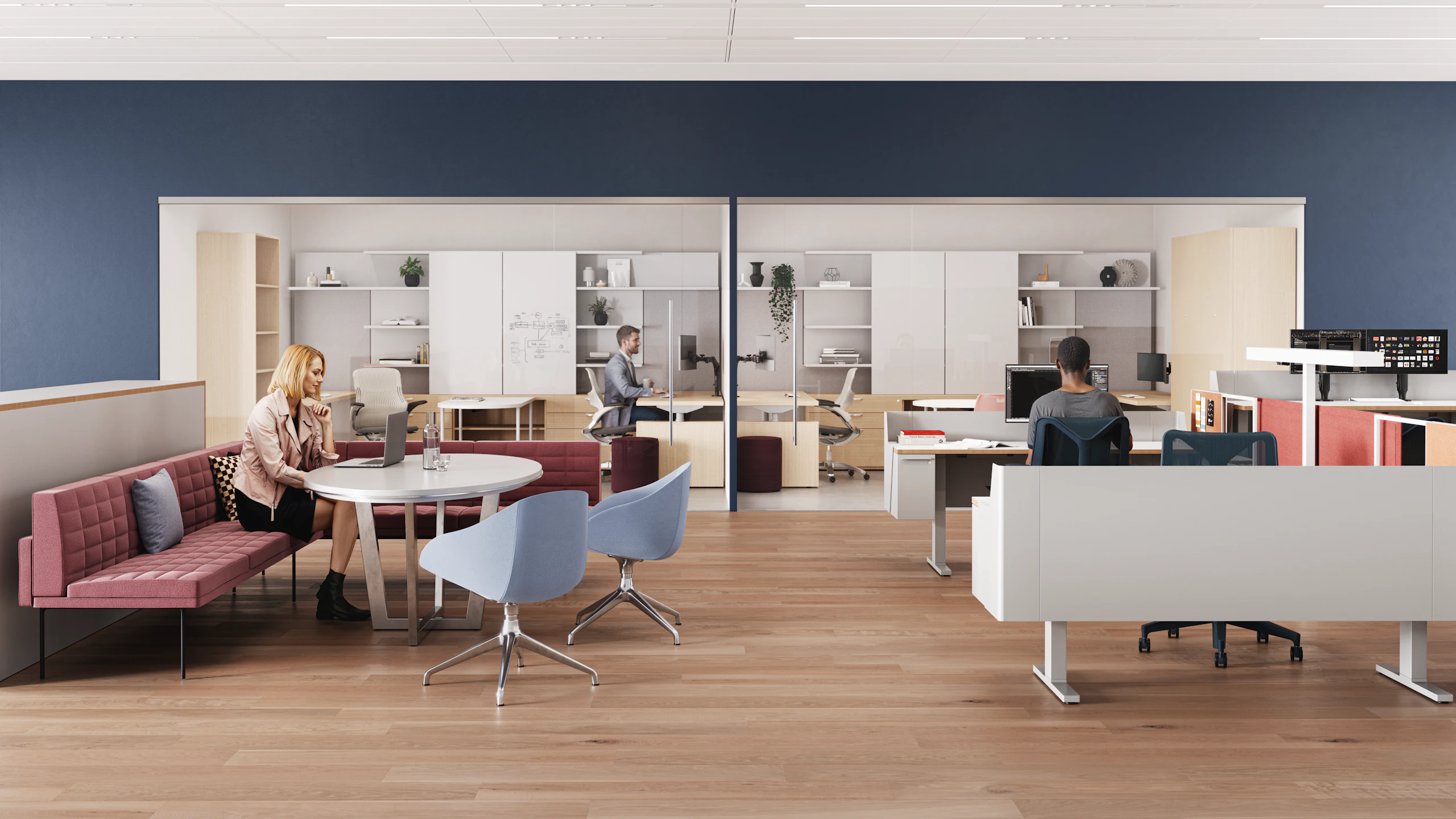 A modern workspace with wooden flooring, featuring an individual seated at a round table, along with a person working at a desk behind glass, and contemporary furniture, including red and blue chairs.