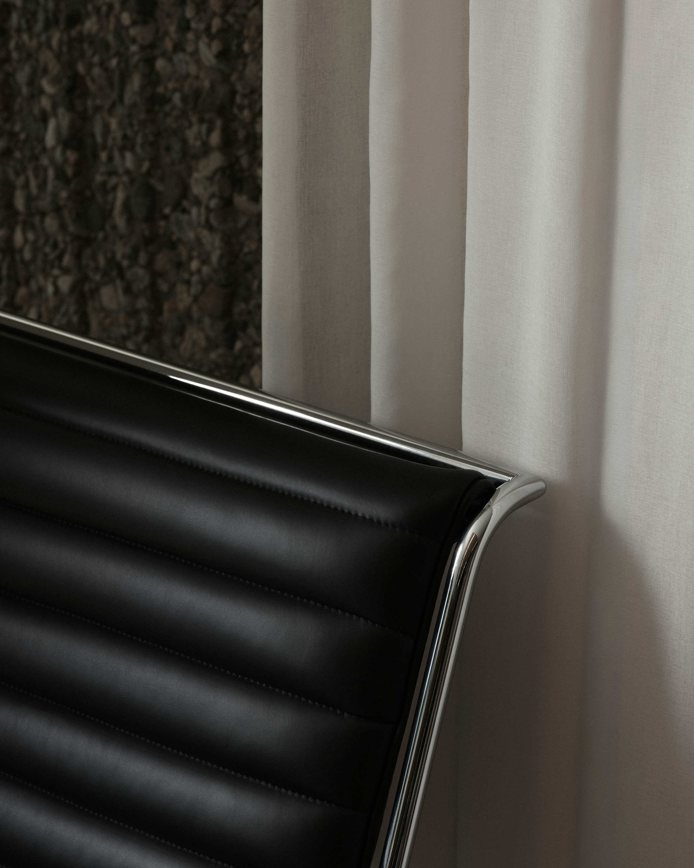 Detail view of the top corner of a Sled Chair backrest, upholstered with black leather with a chrome frame, viewed at an angle against a white curtain and stone wall.