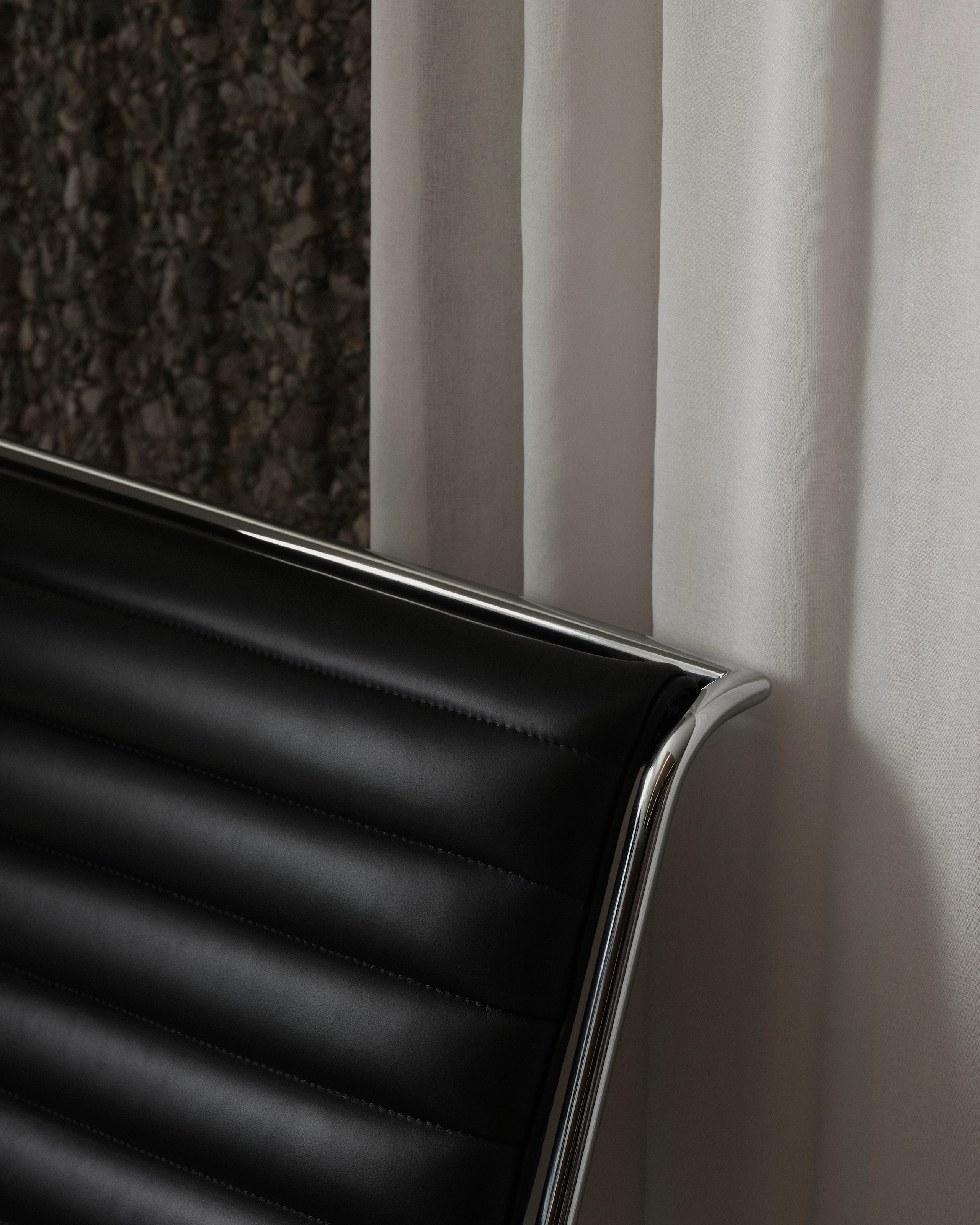 Detail view of the top corner of a Sled Chair backrest, upholstered with black leather with a chrome frame, viewed at an angle against a white curtain and stone wall.