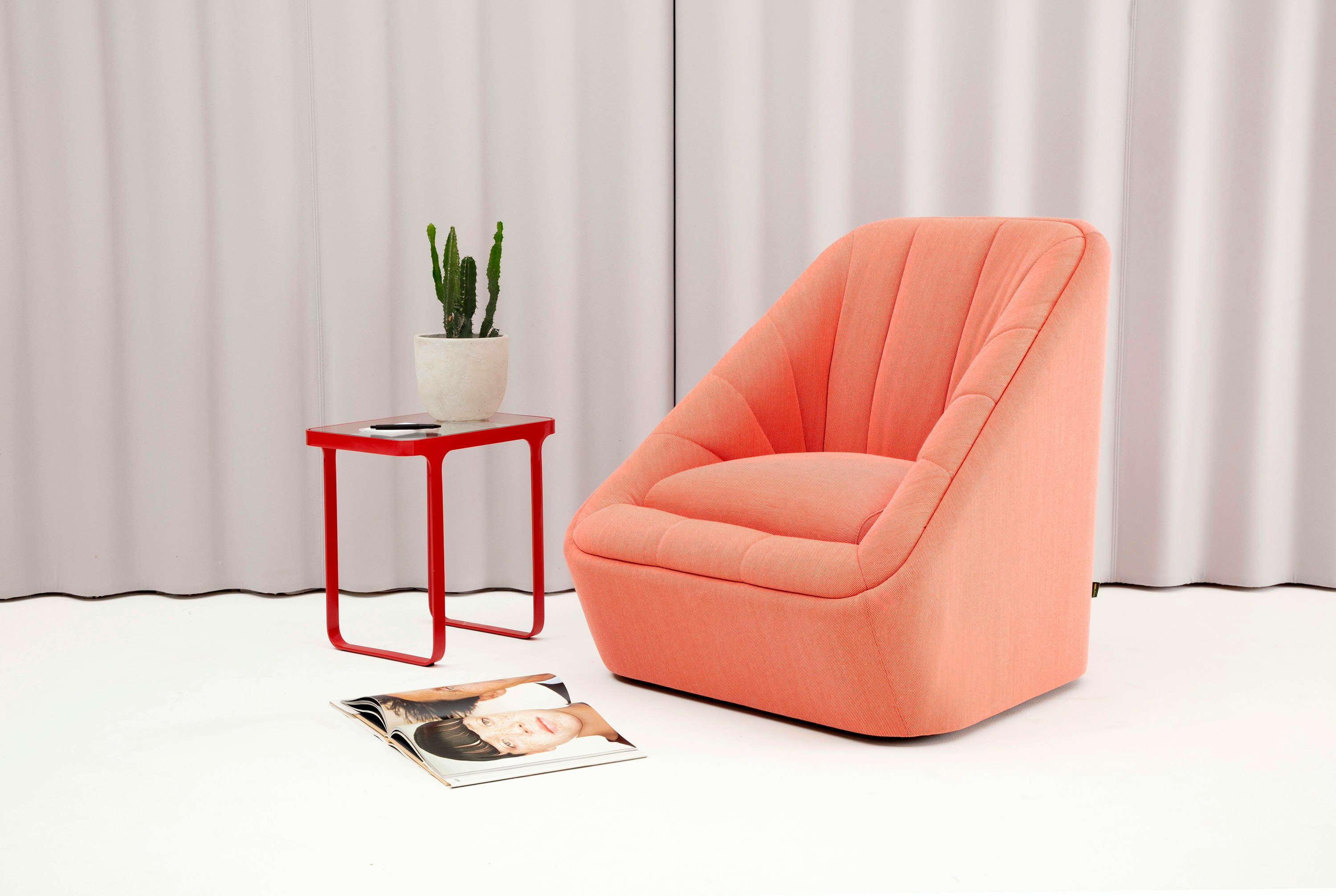 A red naughtone Trace Side Table with glass top alongside a coral Fiji Chair with a curtain backdrop.