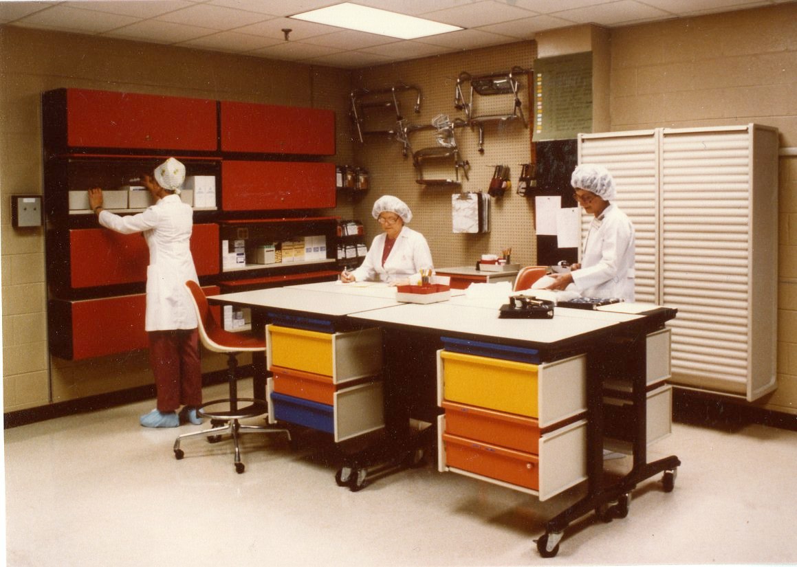  Archival photo of three women working in a room full of colorful cabinets, desks, tables, and stools. 