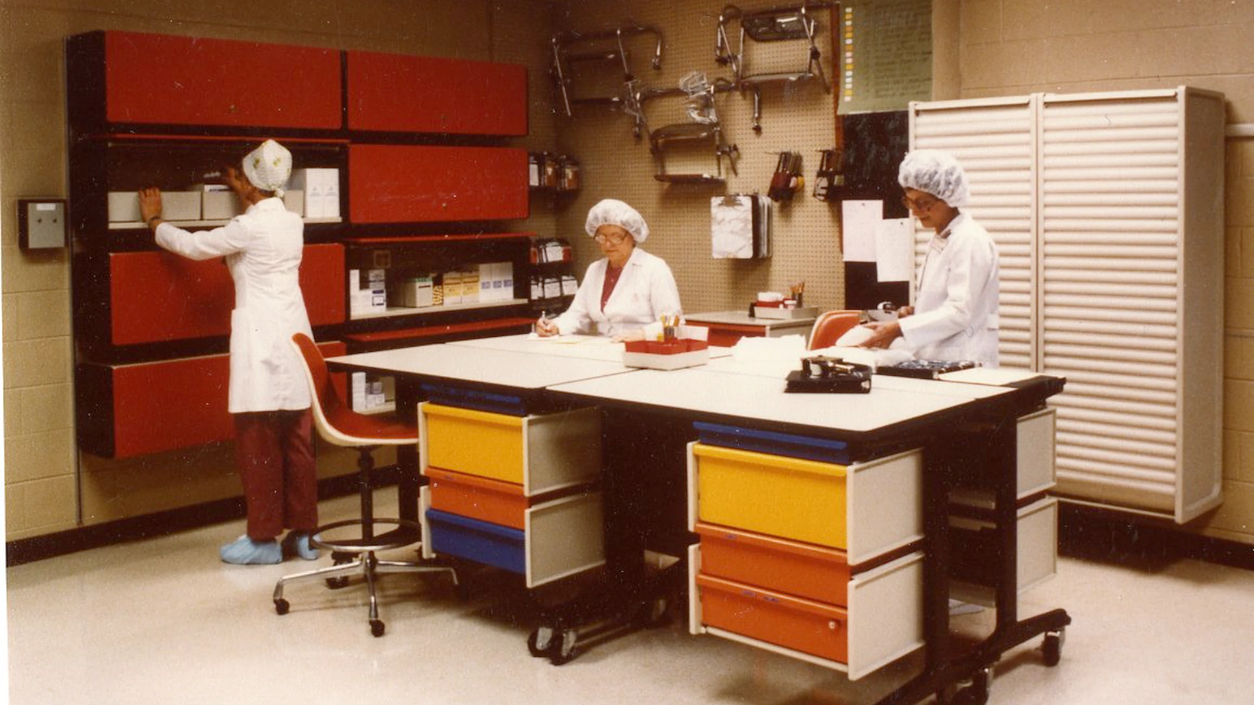 Archival photo of three women working in a room full of colorful cabinets, desks, tables, and stools.